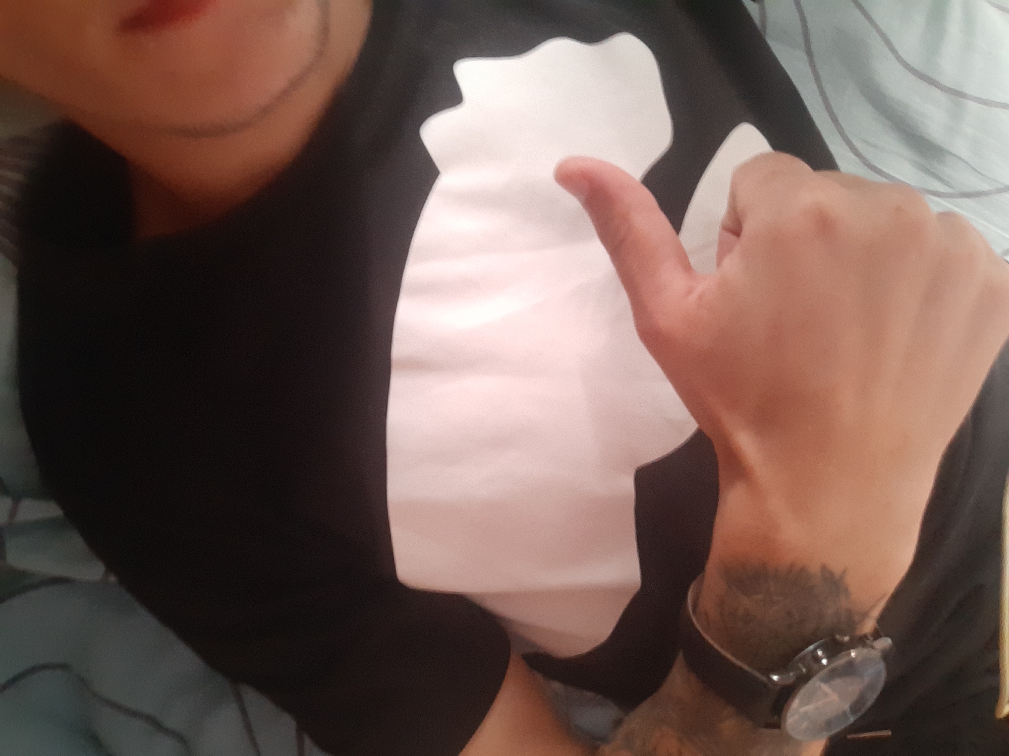 The image shows a close-up, top-down view of a person lying down, with their head and upper body filling most of the frame. The person is wearing a black long-sleeved shirt with a large white abstract shape on the front. Their left arm is visible, with a watch on their wrist and tattoos on their forearm. The person's right hand is extended, with a finger pointing downwards towards the white shape on their shirt. The background appears to be a bed with light blue and white striped sheets. The scene is indoors, and the lighting is soft and diffused. There is no clear indication of the location within Cricova, Moldova, as the image focuses solely on the individual and their immediate surroundings.