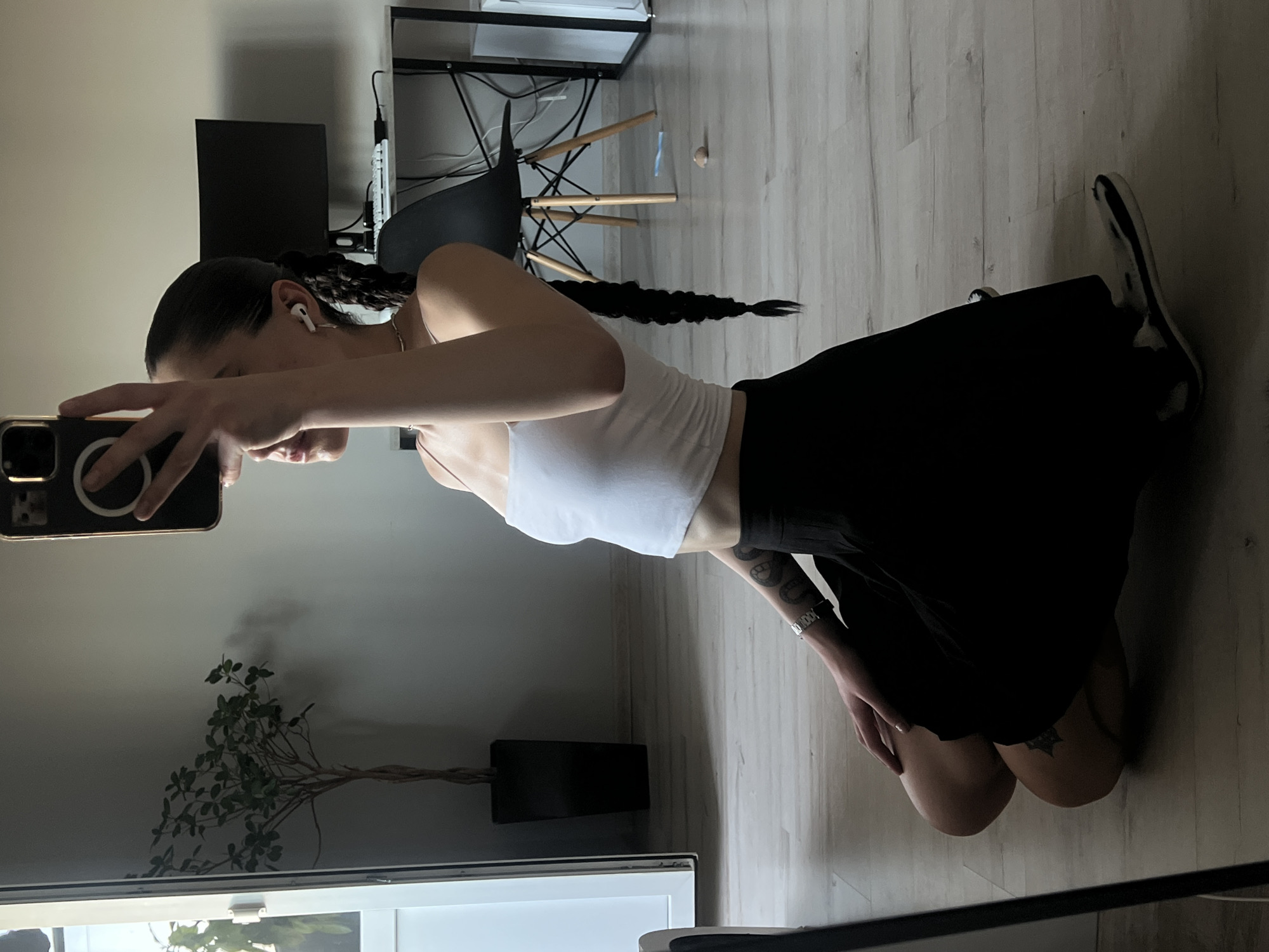 A person kneels on a wooden floor, holding a smartphone to take a picture. They are wearing a white crop top, a black skirt, and white sneakers. Their hair is pulled back into a long braid. The person has tattoos on their arm and thigh. A plant is visible in a black pot next to them. In the background, there is a desk with a computer monitor and a chair. The room appears to be an indoor setting, possibly a home office or bedroom, with neutral-colored walls and natural lighting. The time of day is not specified, but the lighting suggests it is daytime. There is no visible text in the image.