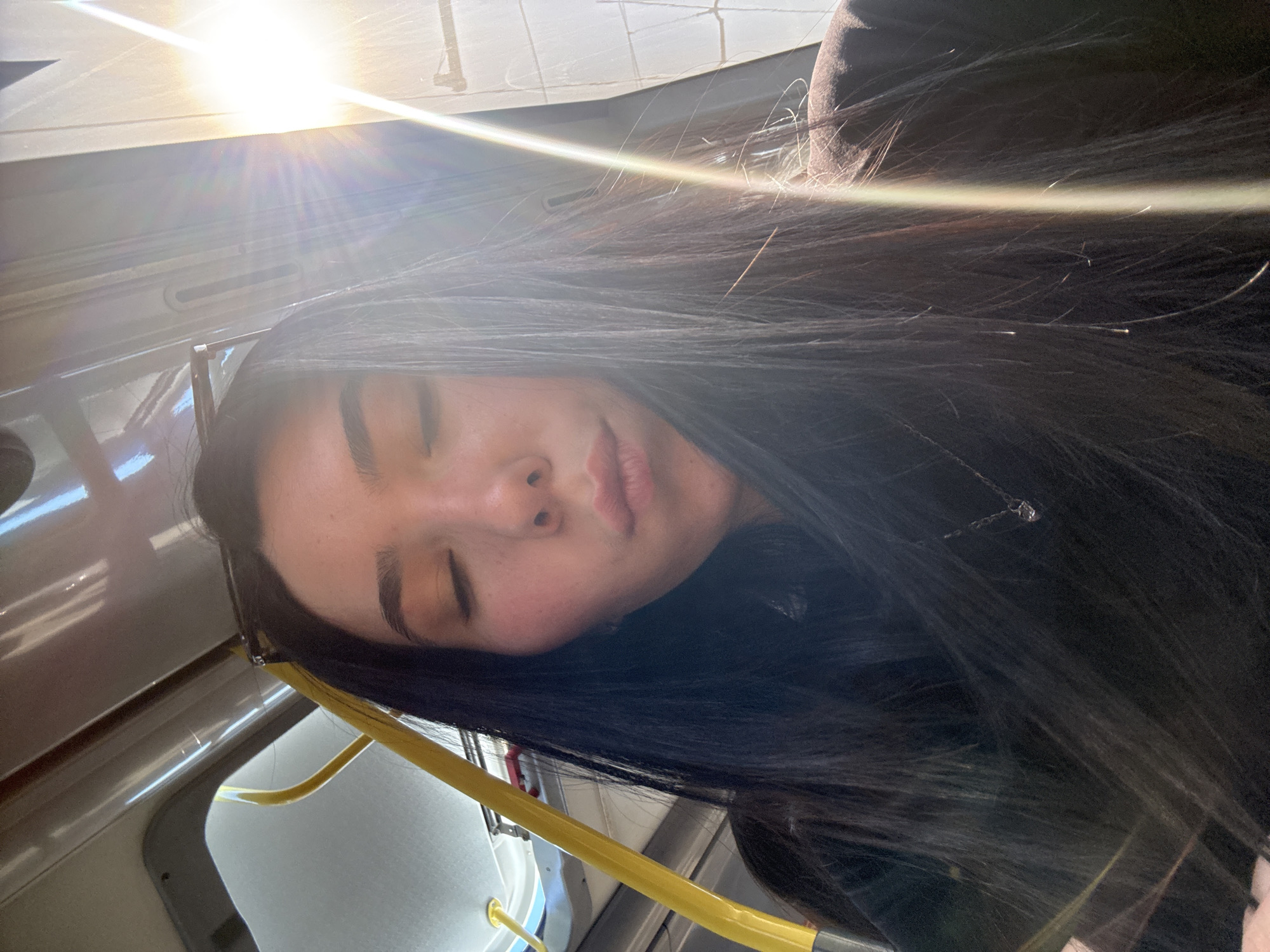 A young woman with dark hair is asleep on public transportation, likely a bus or tram. She is leaning against a window and has glasses next to her face. A yellow handrail is visible. The photo appears to have been taken during the day with bright sunlight. The location is unknown.