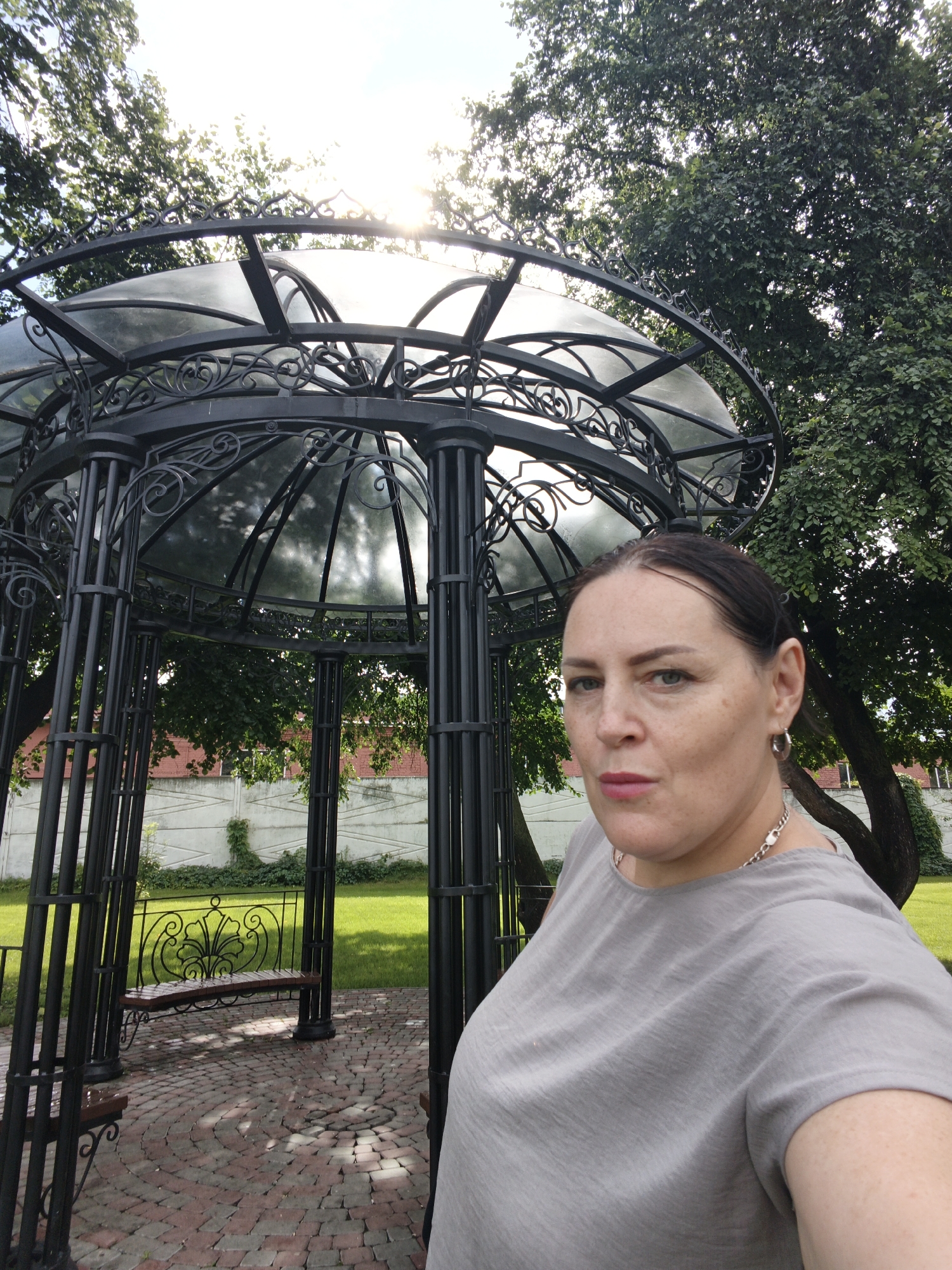 The image shows a woman posing for a selfie in front of a gazebo in the city of Irbit, Russia. The woman is wearing a grey t-shirt and a silver necklace. She has dark hair and light-colored eyes. The gazebo is made of black wrought iron and has a glass roof. It is surrounded by trees and a brick patio. In the background, there is a white fence and a building with a red brick wall. The sun is shining brightly, casting shadows on the ground.
