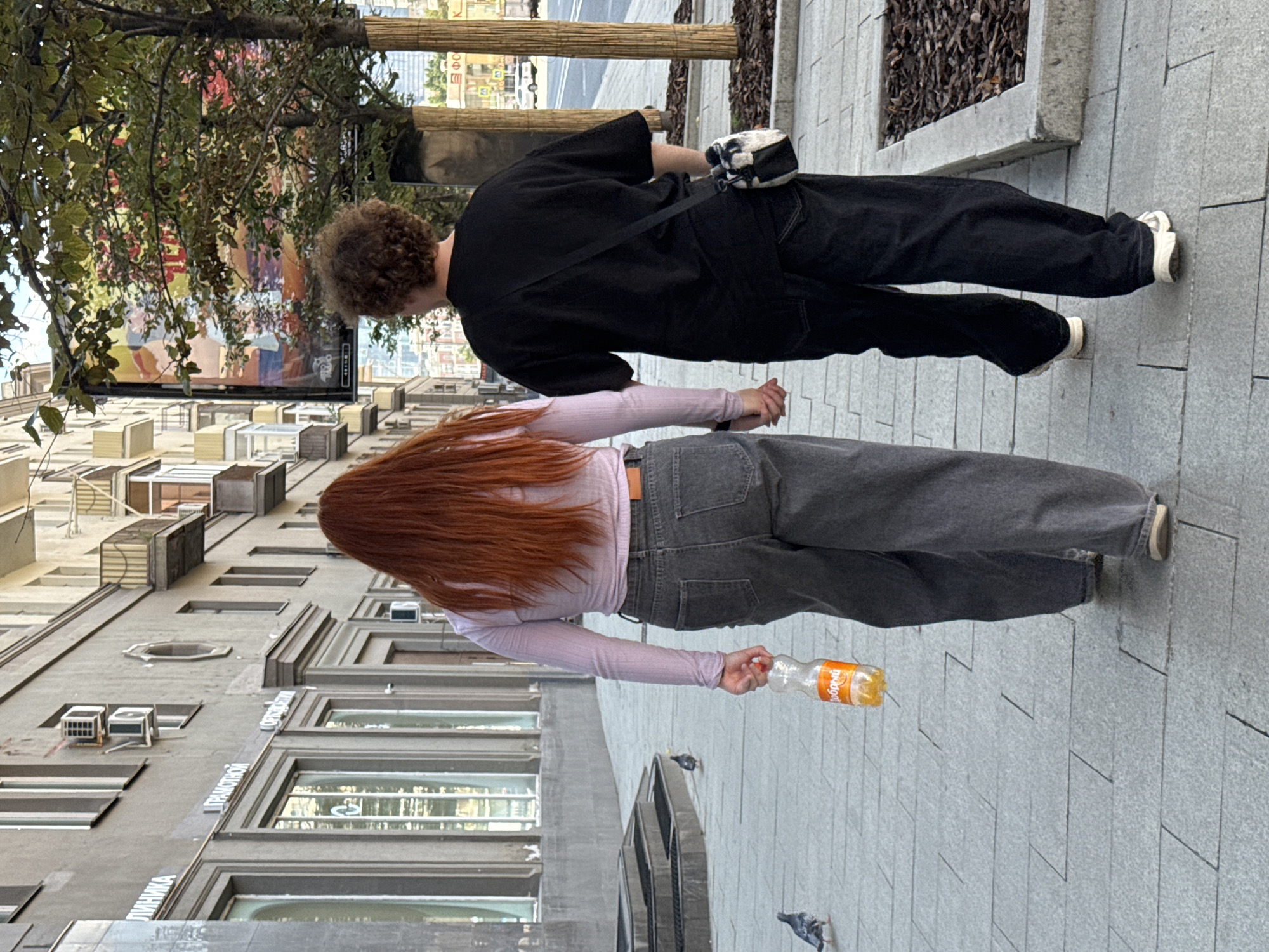 The image shows two individuals walking hand-in-hand on a sidewalk in front of a building. The person on the left has long, reddish-brown hair and is wearing a light purple long-sleeved shirt and grey jeans. They are holding a clear plastic bottle with an orange liquid and an orange label. The person on the right has curly, brown hair and is wearing a black t-shirt, black pants, and white sneakers. They have a small black and white bag slung across their chest. Two pigeons are visible on the sidewalk in the foreground. The building behind them appears to be an older, multi-story structure with windows and balconies. A large advertisement is displayed on the side of the building. The sidewalk is paved with grey rectangular tiles. In the background, there are trees and a street with traffic. The scene suggests an urban environment in Moscow, Russia, based on architectural styles and Cyrillic signage visible on the building.