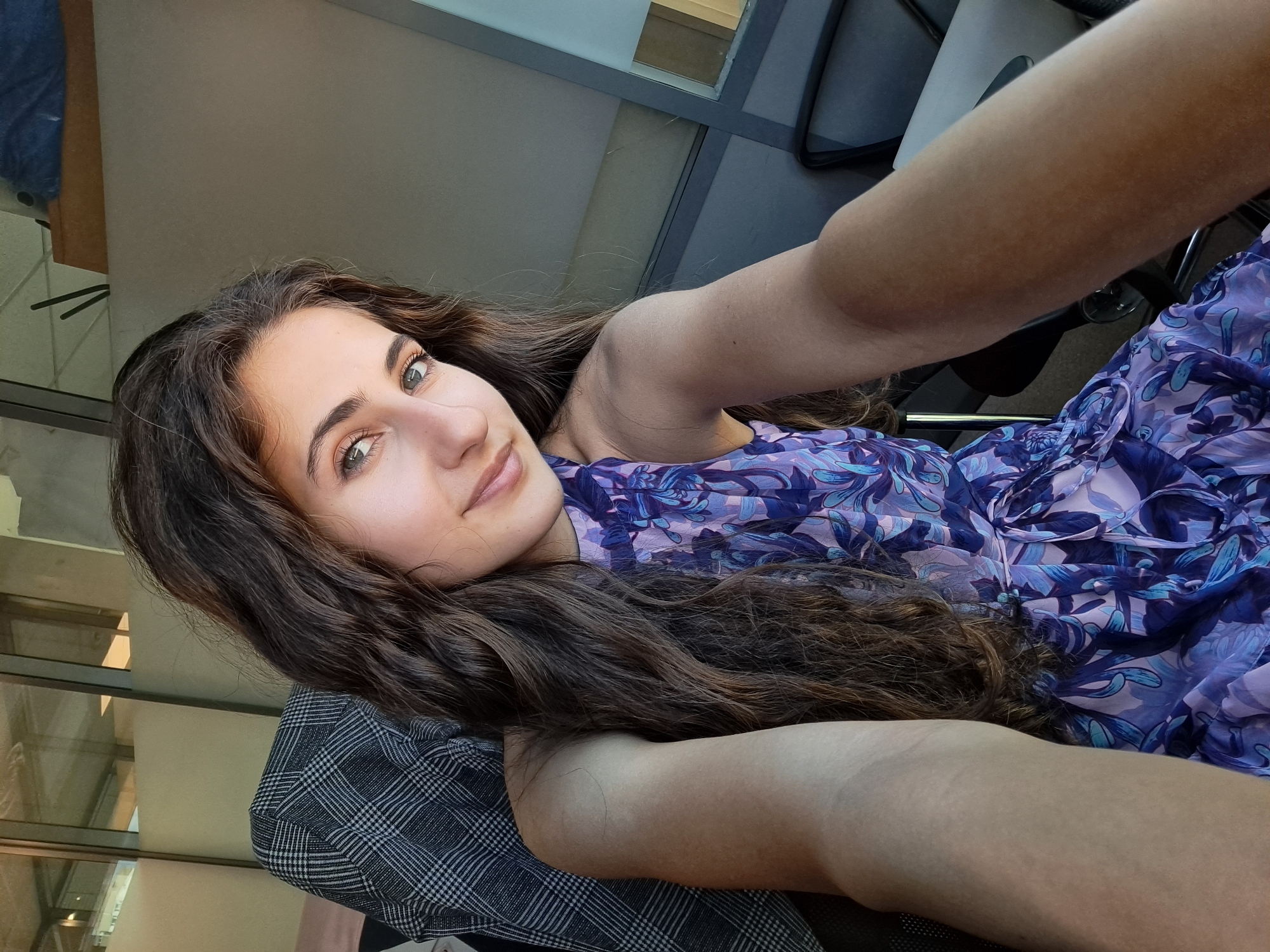 This is a vertically oriented, high-angle, selfie-style photo of a young woman. She has long, wavy, dark brown hair that is draped over her shoulder and chest. Her head is tilted, and she is looking towards the camera with a slight smile. She has striking green eyes and well-defined eyebrows. The woman is wearing a purple floral print dress. In the foreground, her right arm is extended towards the camera, and her left arm is bent at the elbow, resting on her chest. A portion of her torso is visible, clad in a gray and white plaid garment, possibly a jacket or shirt, over the floral dress.
The background appears to be an indoor setting, possibly an office or a cafe, with large windows and reflections visible. The lighting suggests it might be daytime, with natural light filtering in. The overall mood of the image is relaxed and casual. There is no visible text in the image. The location context provided is Sofia, Bulgaria.