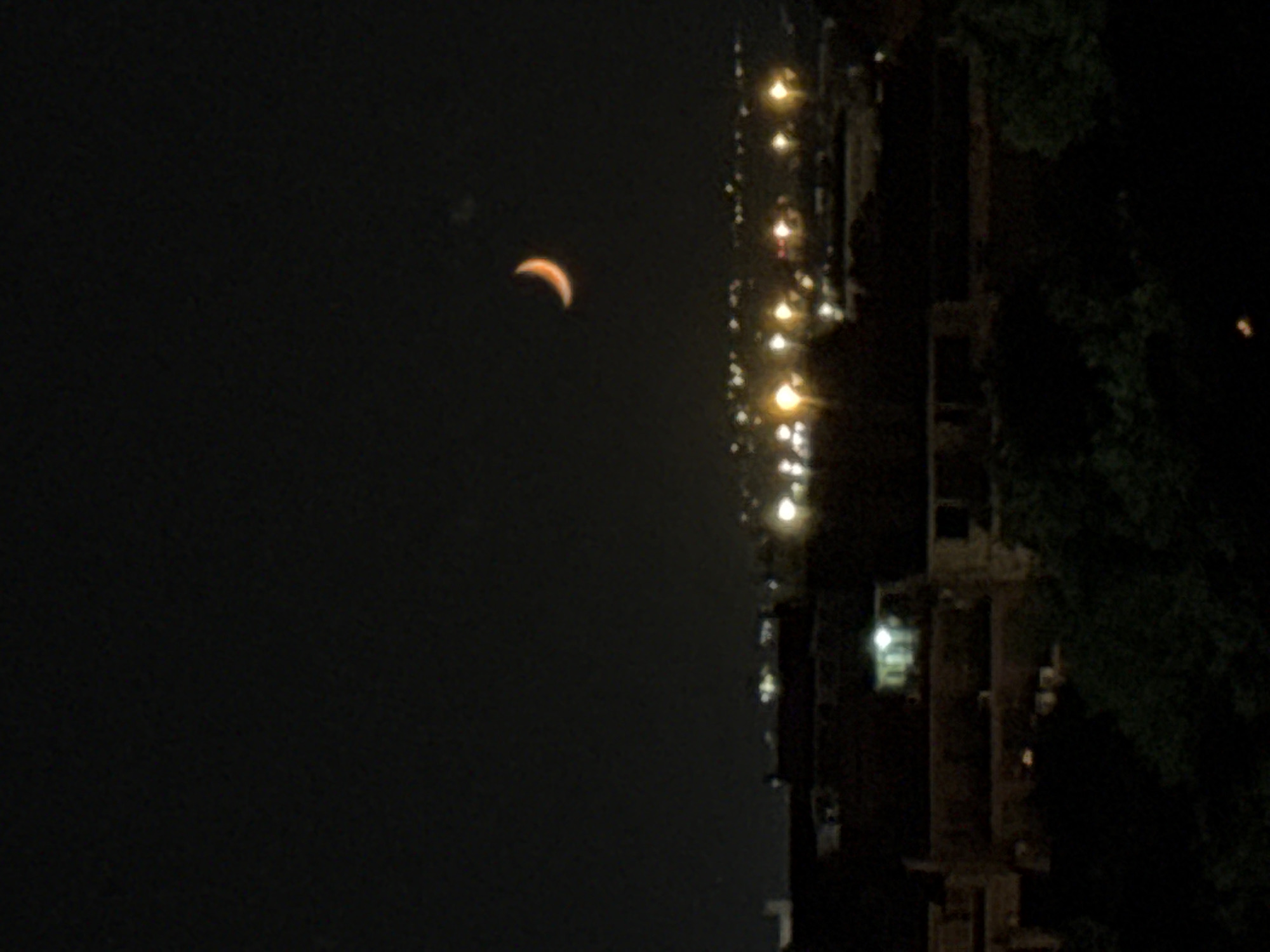 The image, taken at night, features a crescent moon in the upper left portion of the frame. The right side of the image depicts a cityscape, consisting of buildings with illuminated windows and streetlights. Some foliage is visible along the right edge of the buildings. The exact location in Varna, Bulgaria, cannot be determined from the image.