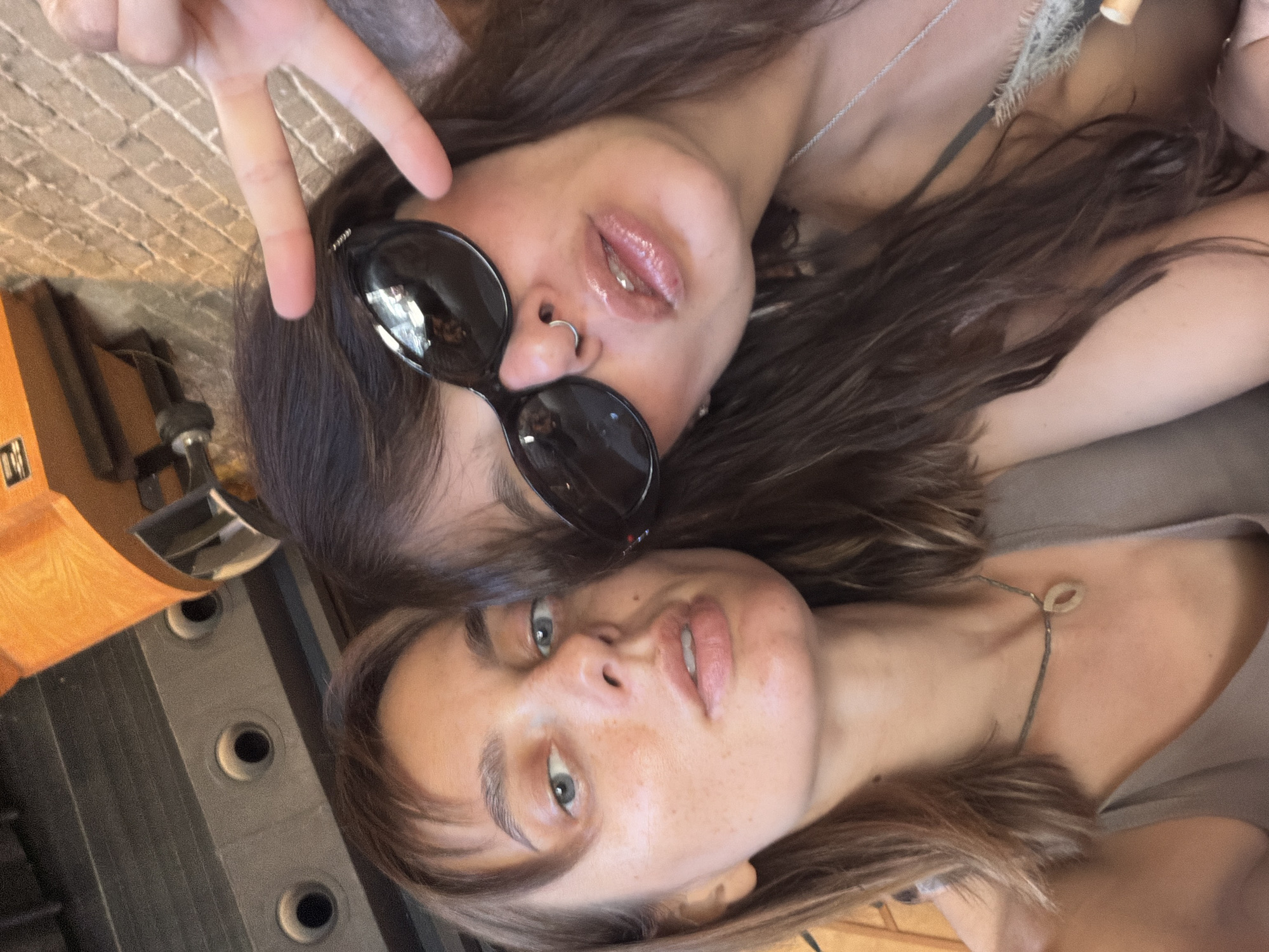 Two young women are taking a selfie in an indoor location. The woman on the left has light brown hair and is wearing a tan top and a necklace. She has freckles on her face. The woman on the right has dark brown hair and is wearing large black sunglasses and a nose ring. She is making a peace sign with her right hand. The background appears to be a restaurant or bar, with brick walls and industrial lighting fixtures visible. The overall atmosphere suggests a casual, social setting.