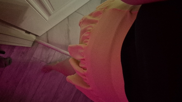 A low-angle shot captures a person in a yellow garment with ruffles, possibly a dress or top, contrasted by a black garment. A bare foot is visible on a floor with pink lighting. In the background, a white door or cabinet is slightly ajar. The lighting creates a moody atmosphere.