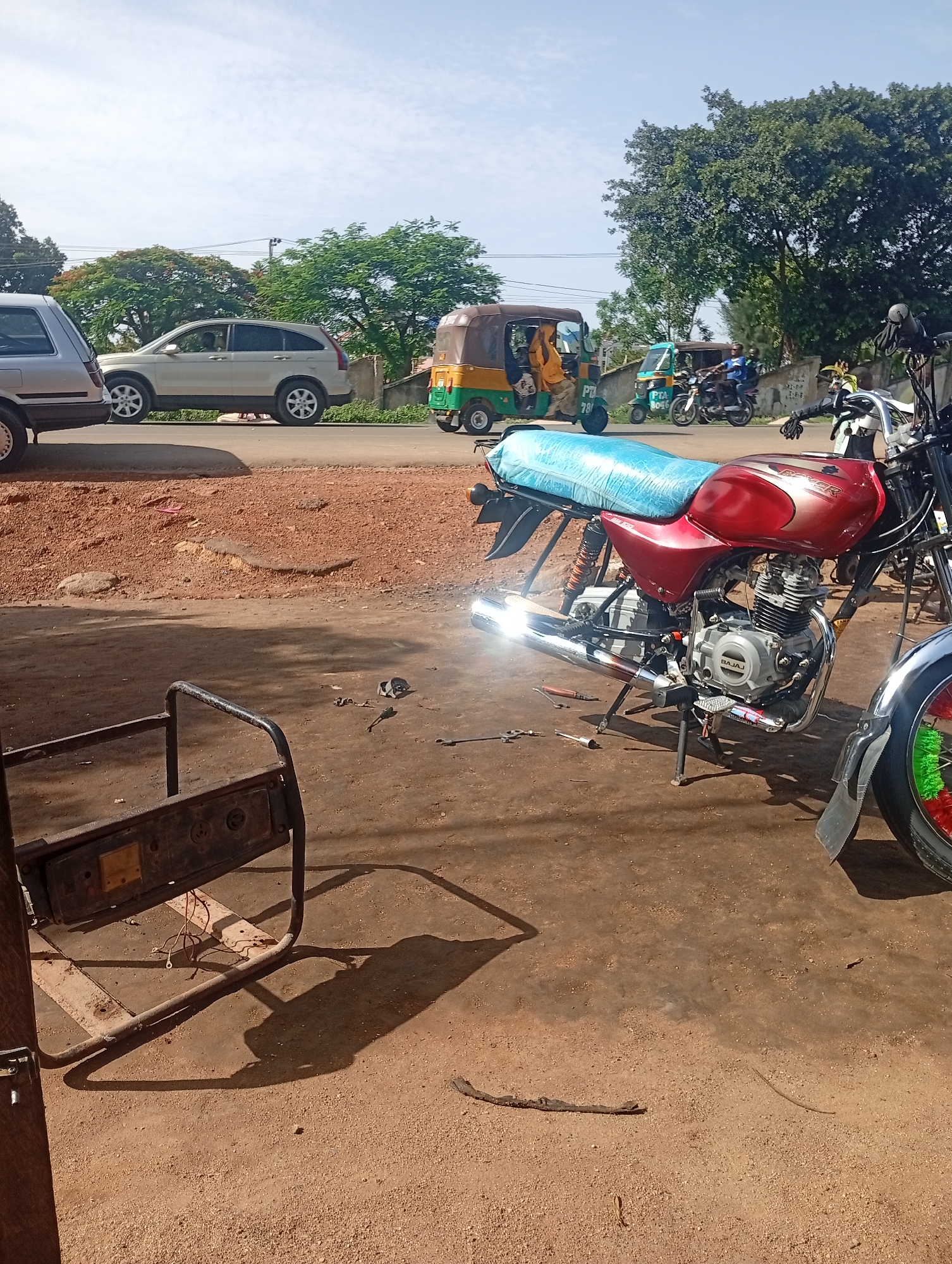 In Rafin Par, Nigeria, on a dusty, sun-drenched day, a street scene unfolds with vehicles and a motorcycle under repair. The foreground features the partially disassembled frame of a generator with wires dangling, resting on the sandy ground. Tools, including pliers and a screwdriver, are scattered nearby, indicating ongoing work. A red Bajaj Boxer motorcycle, covered with a light blue tarp, is the central focus. Its engine and exhaust are exposed, suggesting mechanical attention. To the right, the rear wheel of another motorcycle, adorned with colorful green and red trim, is visible. In the mid-ground, a beige SUV is parked on a paved road, and further down the road, a yellow and green auto-rickshaw carries passengers. Beyond the road, lush green trees and a wall line the background, with a clear blue sky overhead. The overall impression is a typical roadside scene in a Nigerian town, likely a mechanic's workshop or a busy street corner.