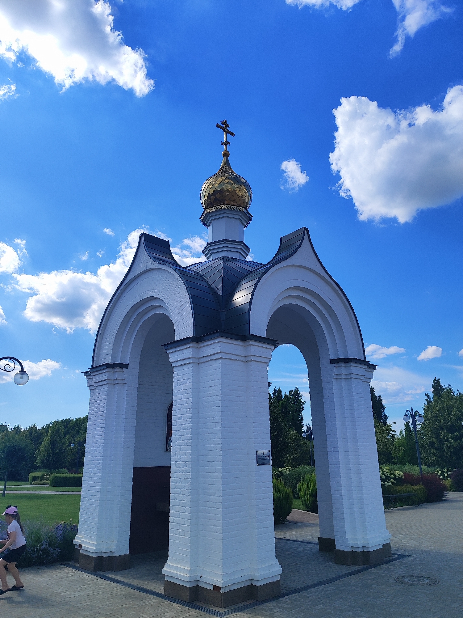 In Bryukhovetskaya, Russia, a white brick chapel with a gilded dome and a cross stands against a bright blue sky dotted with fluffy white clouds. The chapel features four arched entrances and a dark, pointed roof. In the foreground, a young girl with a pink hat and white shirt walks past the chapel. The surrounding environment includes a paved walkway, a well-maintained green lawn, trees, and ornamental bushes. A lamppost with a spherical light fixture is visible on the left side of the image.