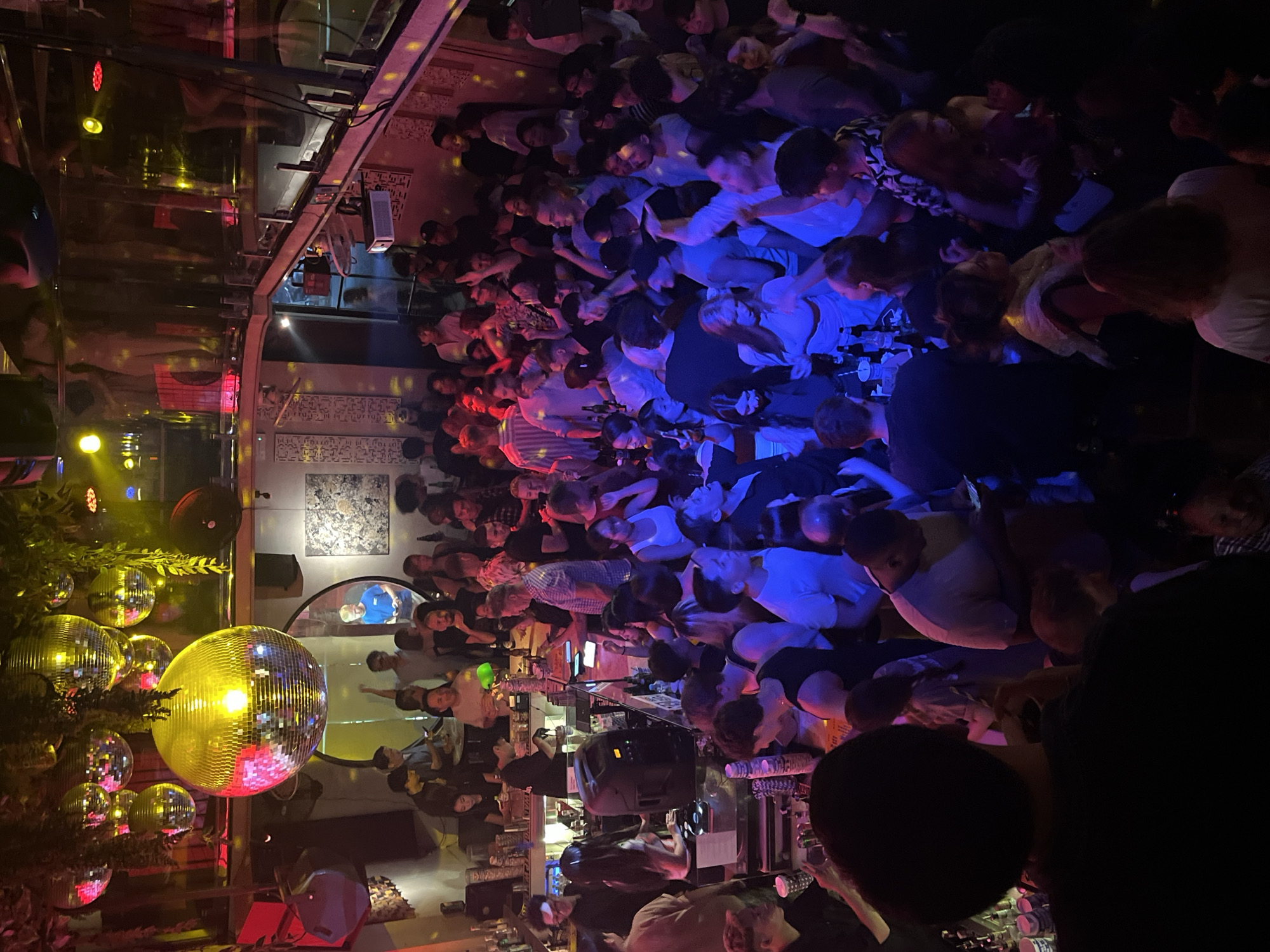 Here's a detailed description of the image:
**General Description:** The image depicts a crowded nightlife scene, likely inside a bar or club, possibly in Đà Nẵng, Vietnam, based on the environment and style. The crowd is densely packed.
**People:** The image shows a large group of people. Their actions include standing, talking, drinking and other associated activities.
**Objects:** The scene includes disco balls, lighting fixtures, bar equipment (bottles, glasses), sound equipment.
**Environment:** The setting appears to be a nightclub or bar environment, indicated by the lighting, music equipment, and the bar setup.
**Additional Notes:** The lighting creates a vibrant atmosphere. The presence of a disco ball, bar, music, and a crowded scene all support the description of this place.