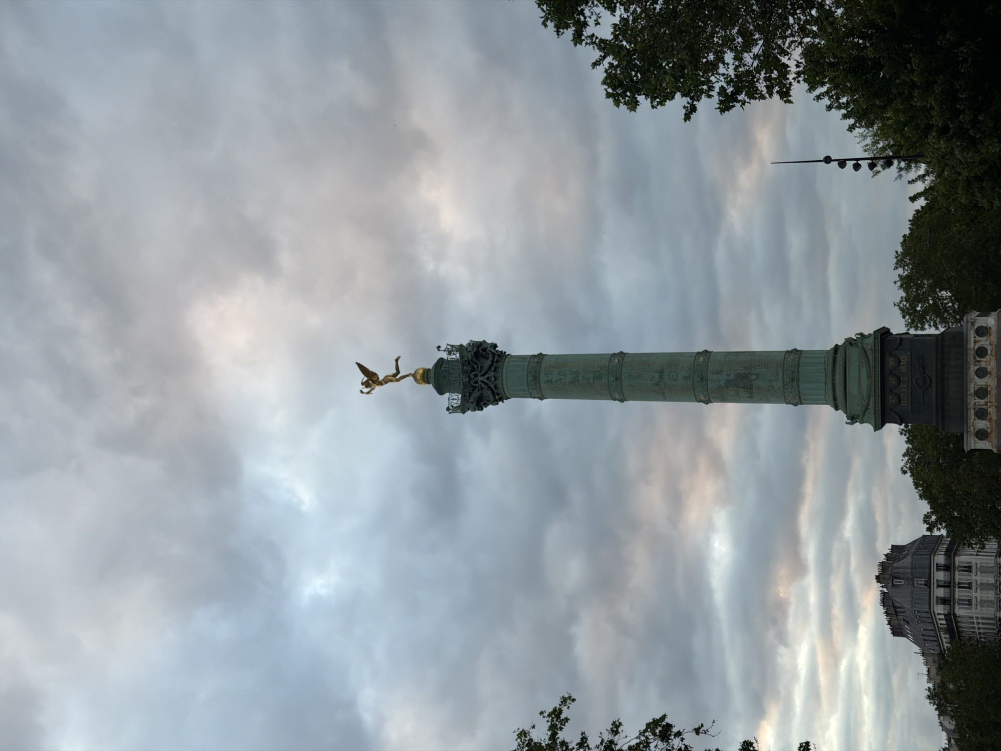 The image shows the Colonne de Juillet (July Column), topped with the golden statue of the "Spirit of Freedom" (Le Génie de la Liberté), situated in the Place de la Bastille in Paris, France. The column is a cylindrical structure with bronze-colored decorations along its surface. The sky is overcast with layered, gray and white clouds. Tree foliage frames the top and bottom edges of the image. A building is visible on the right side of the scene.