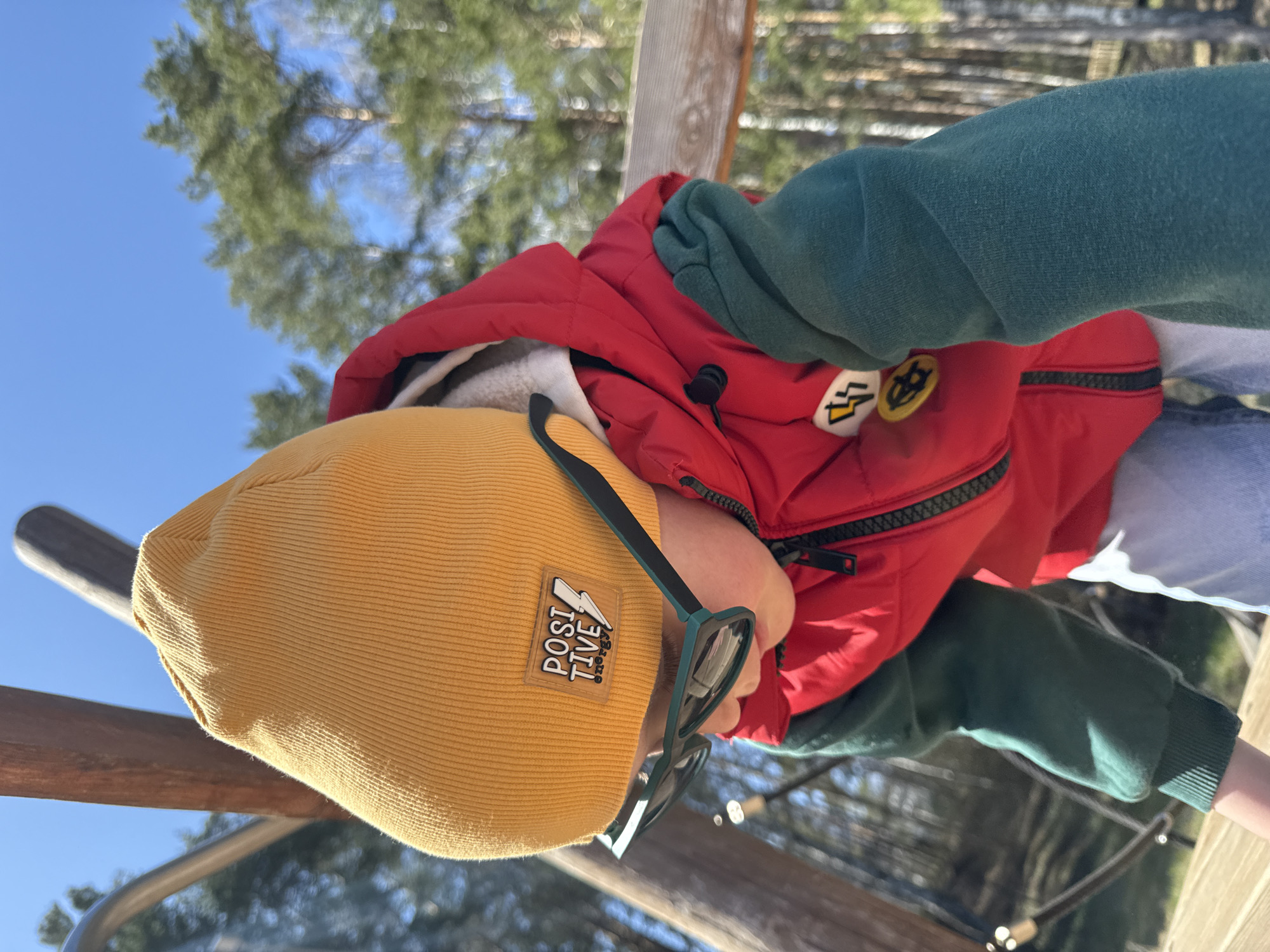 A child wearing a yellow beanie with a "Positive Energy" patch, a red jacket, and green sleeves is standing on a wooden structure. The child also wears eyeglasses. The sky is blue and there are trees in the background. The location is likely Chaikovsky, Russia.