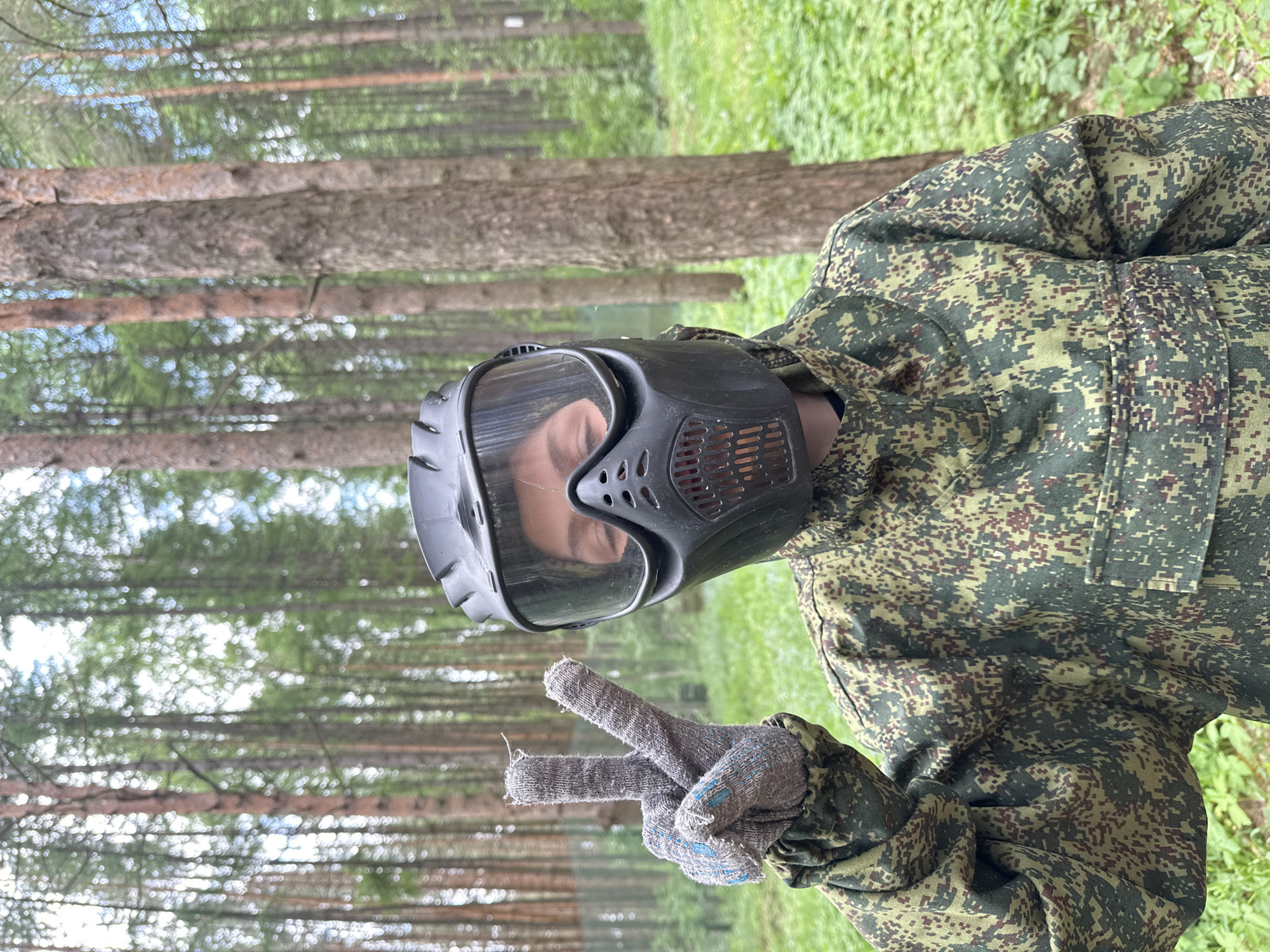 A person wearing camouflage clothing and a full-face paintball mask is standing in a forest and making a peace sign. The person's gloved hand is visible. Trees are in the background, as is some green foliage. The location is consistent with the forests around Votkinsk, Russia.