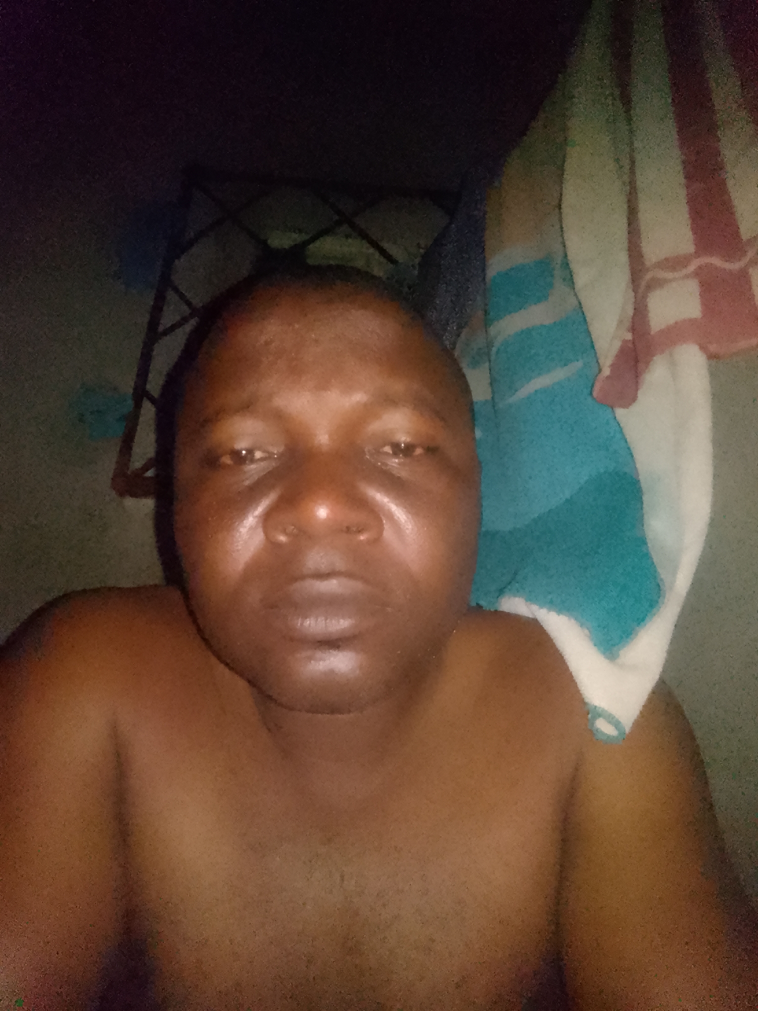 A shirtless person with dark skin is facing the camera, their face slightly shiny. Behind them, there's a dark metal rack or shelf. To the right, a striped towel hangs, showing white, teal, and pink sections. The background is dimly lit and appears to be a room with plain walls. The location is Unknown, Unknown.