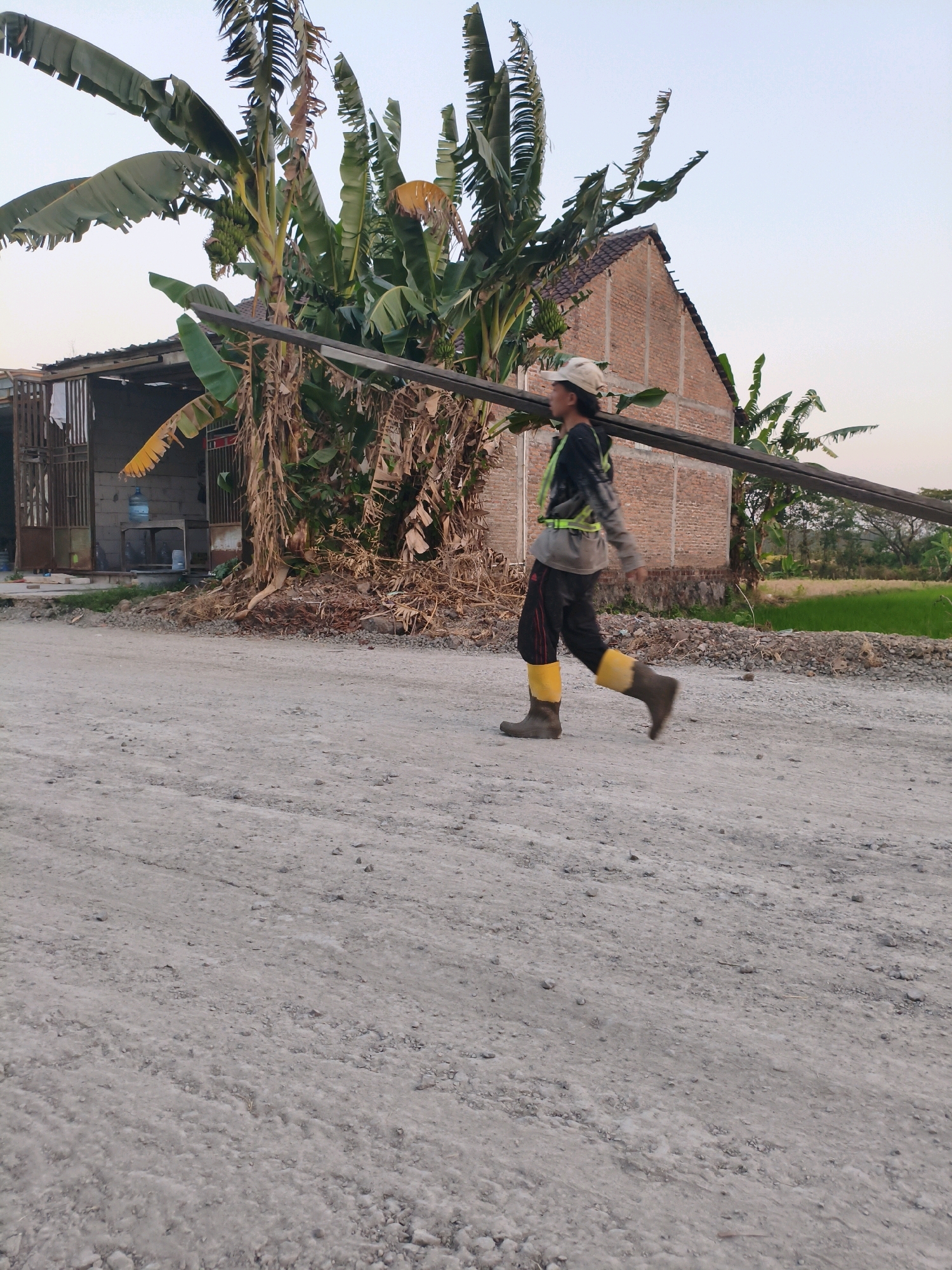 In the city of Gondangrejo, Indonesia, a male construction worker wearing a light-colored cap, a black and gray long-sleeved shirt with a reflective vest, dark pants, and yellow and brown rubber boots walks from left to right across a dirt road. He is carrying a long, dark metal bar horizontally across his shoulders. Behind him, a building constructed of brick with a tiled roof is partially visible, along with several banana trees. The ground is covered in gravel and dirt. The sky above is clear.