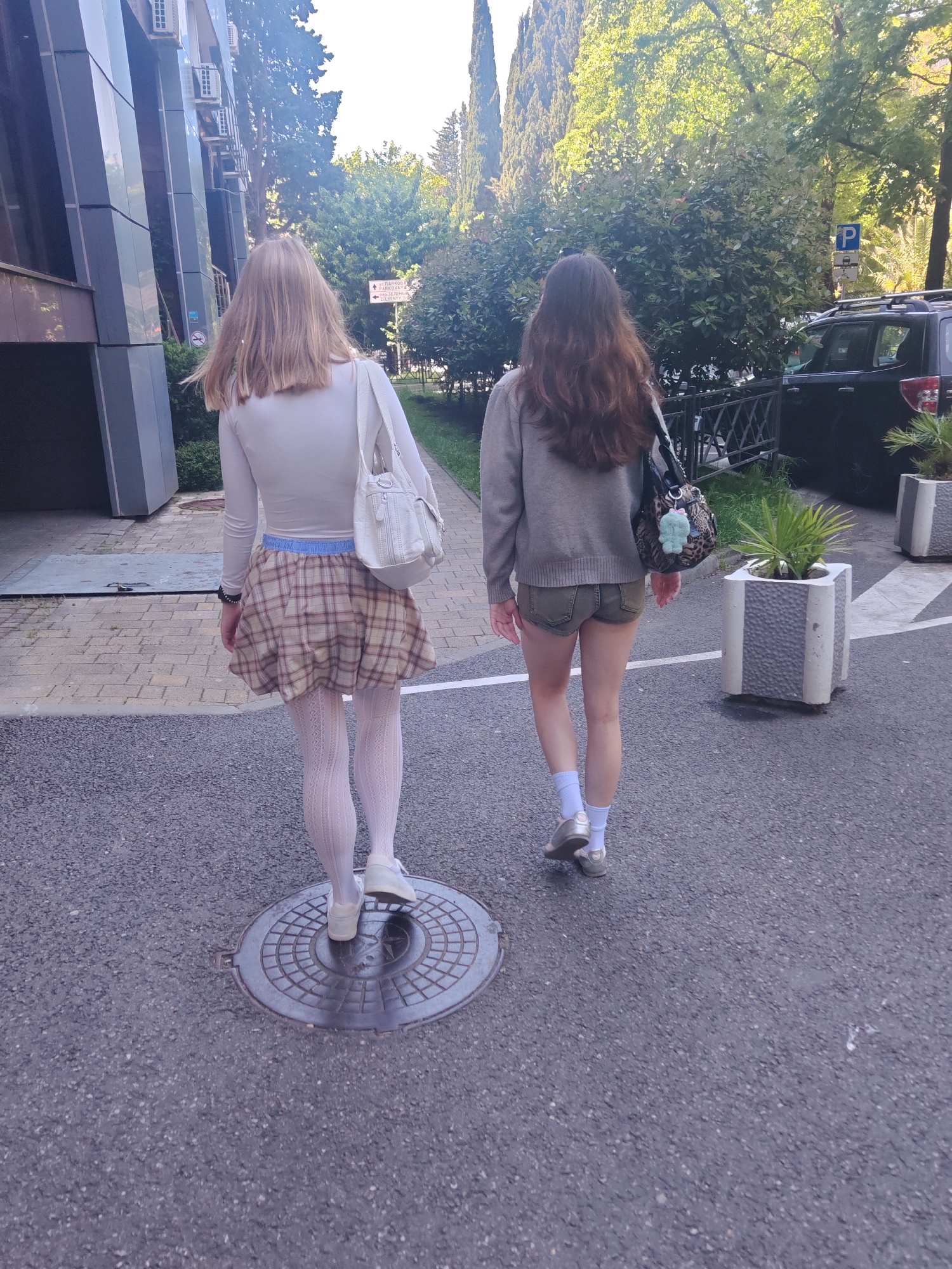 Two young women walk away from the viewer on an asphalt road next to a sidewalk and a building. The scene appears to be an urban or suburban street on a bright, sunny day.
The person on the left has light brown or blonde hair, wears a white long-sleeved top, a beige and brown plaid mini-skirt with a light blue elastic waistband, white patterned tights, and white sneakers. She carries a white shoulder bag on her left shoulder. She is stepping onto a circular metal manhole cover in the foreground.
The person on the right has long dark brown hair, wears a grey long-sleeved top or sweater, olive green shorts, white ankle socks, and silver or grey shoes. She carries a black shoulder bag with a leopard print pattern and a light blue/green fluffy keychain.
To the left, a modern building with dark reflective panels and multiple white air conditioning units is visible. To the right, there's a lush green landscape with trees, some tall and slender, and general foliage. A black SUV-type car is parked in the distance behind a metal fence. Concrete planters with small palm-like plants line the road.
Notable details include a blue parking sign with a white 'P' symbol and additional symbols indicating parking rules. A white rectangular street sign reads "ПАРКОВАЯ" (PARKOVA), "UL. GAGARINA", and "SEVERNYY", indicating a Russian-speaking location. The bright sunlight casts distinct shadows, suggesting it is either morning or afternoon. No specific emotions are discernible as the subjects have their backs to the camera.