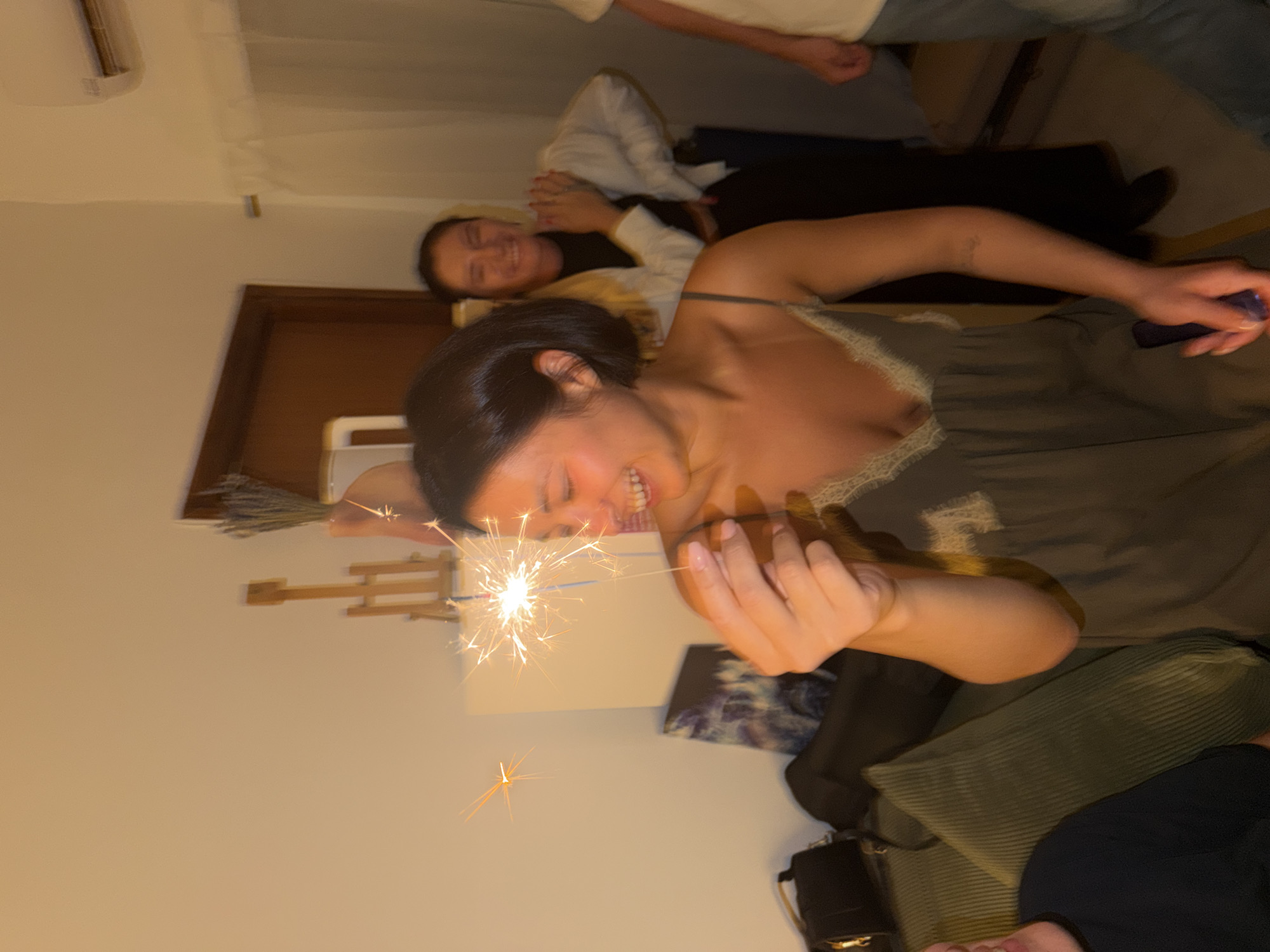 A medium shot captures a woman in the foreground, holding a lit sparkler in her right hand. She is smiling broadly, with her head tilted to the right, and her eyes are closed. Her dark hair is short and styled, falling just above her shoulders. She is wearing a dark, spaghetti-strap dress with a lace trim along the neckline. In the background, two other individuals are visible. To the right of the woman in the foreground, another woman with dark hair is smiling and looking towards the camera, wearing a white jacket over a dark top. Further to the right, a person's torso is visible, wearing a light-colored t-shirt and blue jeans. The environment appears to be an indoor setting, possibly a living room, with a couch in the lower left corner of the frame. There is a wall behind the subjects, and a glimpse of a door and some decorative items on a shelf is visible. The lighting is warm and somewhat dim, with the sparkler being the brightest light source. The image does not provide any specific visual cues to identify its location as Tel-Aviv, Israel.