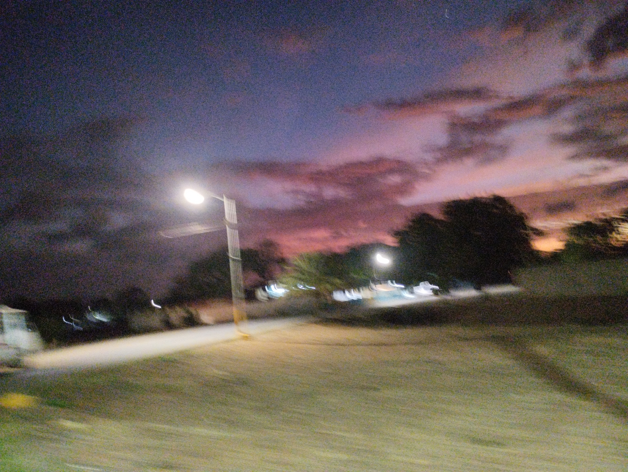 This is a blurry, nighttime photograph taken in the city of La Guardia, Venezuela. The image captures a street scene with a prominent street light on the left side, illuminating a paved area and a grassy section. In the background, the sky is a gradient of dark blue transitioning to shades of purple and pink, suggesting a twilight or early evening sky with some cloud cover. Silhouetted trees are visible against the colorful sky. There are some blurry lights in the distance, possibly from other street lights or vehicles. The overall impression is of a quiet, outdoor location at dusk.