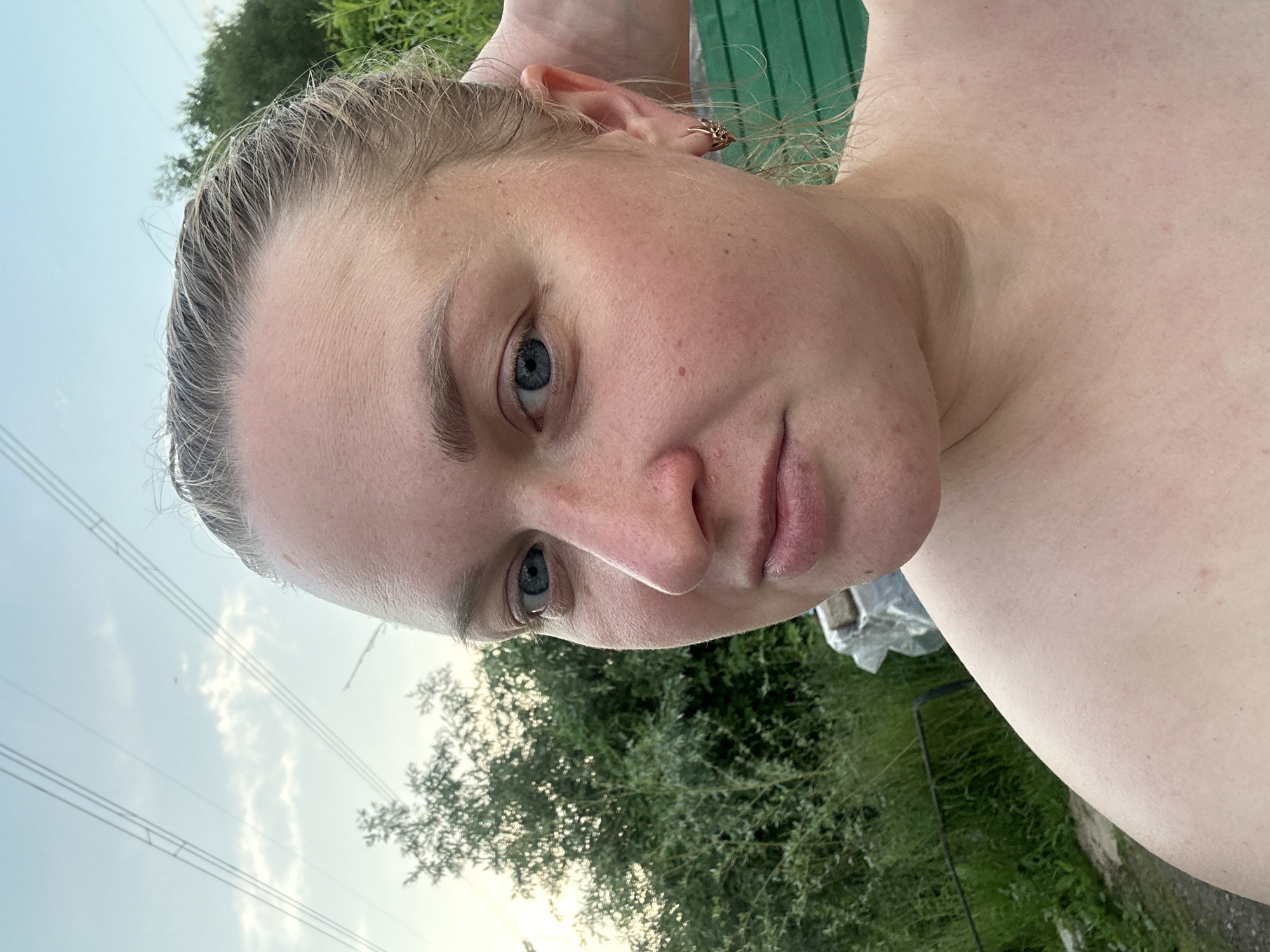 A selfie of a fair-skinned woman with blonde hair pulled back is taken outdoors, likely in a rural setting. She is looking towards the camera with a slight smile. Her right hand is resting on the back of her head, revealing a small, gold-colored hoop earring in her right ear. The background shows lush green foliage and a pale blue sky with wispy clouds. Overhead, power lines stretch across the sky. A glimpse of a green wooden structure is visible on the right side of the frame. The overall scene suggests a casual, outdoor moment.