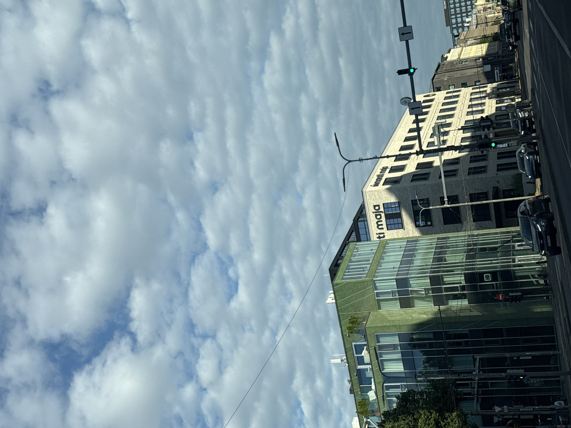 This image captures a street view in Tallinn, Estonia, featuring a modern green building with a glass facade on the left, alongside other multi-story buildings, some with signage. A street lamp with two heads is visible in the mid-ground. Traffic lights are present, with one displaying a green light. Cars are on the road, and a pedestrian signal is also visible. The sky is partly cloudy with a bright, overcast appearance. The scene appears to be captured from a moving vehicle on a city street.