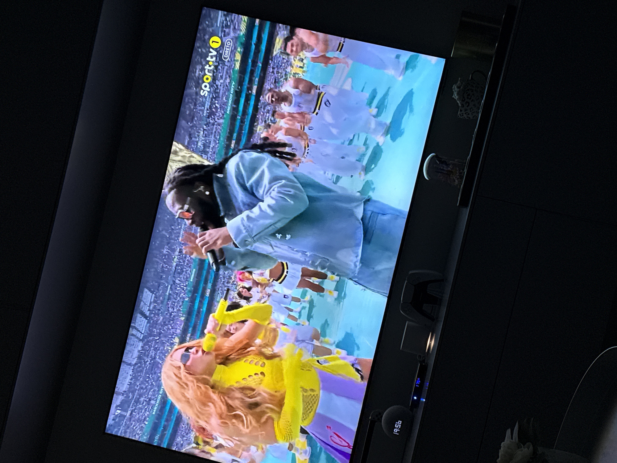 The image shows a television screen displaying a live performance. On the screen, a female performer with long, blonde hair and a yellow outfit is singing into a yellow microphone. She is wearing sunglasses. Next to her, a male performer with dark hair and a light blue jacket is also holding a microphone. They are on a stage with a large crowd in the background. The "sport.tv 1" logo and the word "DIRETO" are visible in the upper right corner of the screen, indicating it is a live sports broadcast. Below the TV screen, on a dark surface, are several objects including a digital clock showing "19:56", a game controller, and a coffee mug. The scene is dimly lit, with the light from the television illuminating the surrounding objects. There is no indication of the city of Differdange, Luxembourg in this image.