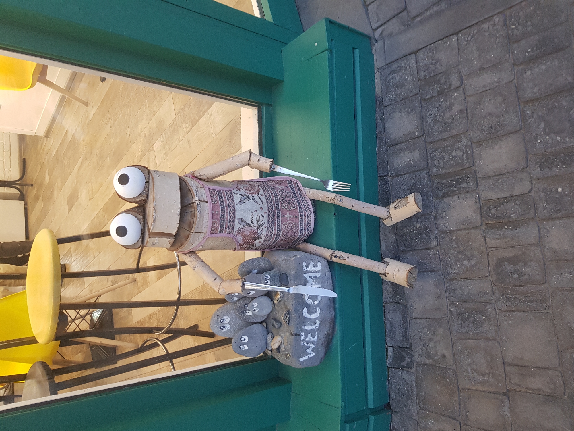 This is a full, eye-level, outdoor shot of a wooden figurine with googly eyes, wearing an apron, sitting on a green ledge in front of a window display, in the city of Unknown, Unknown. The figurine is made of what appears to be birch logs for its body and limbs. It has large googly eyes on top of its head. The figurine is holding a knife in its right hand and a fork in its left. To its left, a large grey rock with "WELCOME" painted on it is visible, with several smaller painted rocks with faces on them sitting on top of it. The ledge it is sitting on is green. The background shows a reflection of yellow stools and tables inside a store. The ground in front of the ledge is paved with grey cobblestones. The scene is brightly lit, suggesting it is daytime.