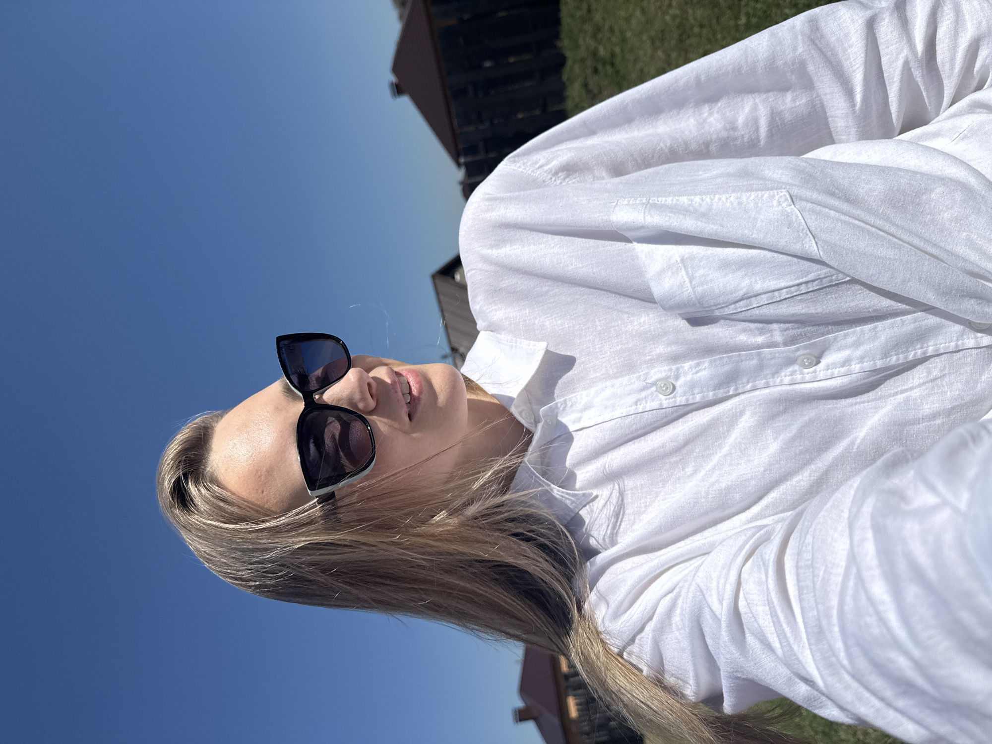A woman with long, blond hair is wearing dark sunglasses and a white button-down shirt. The background is a bright blue sky. The location is Chaikovsky, Russia. A structure of some kind is visible in the background.