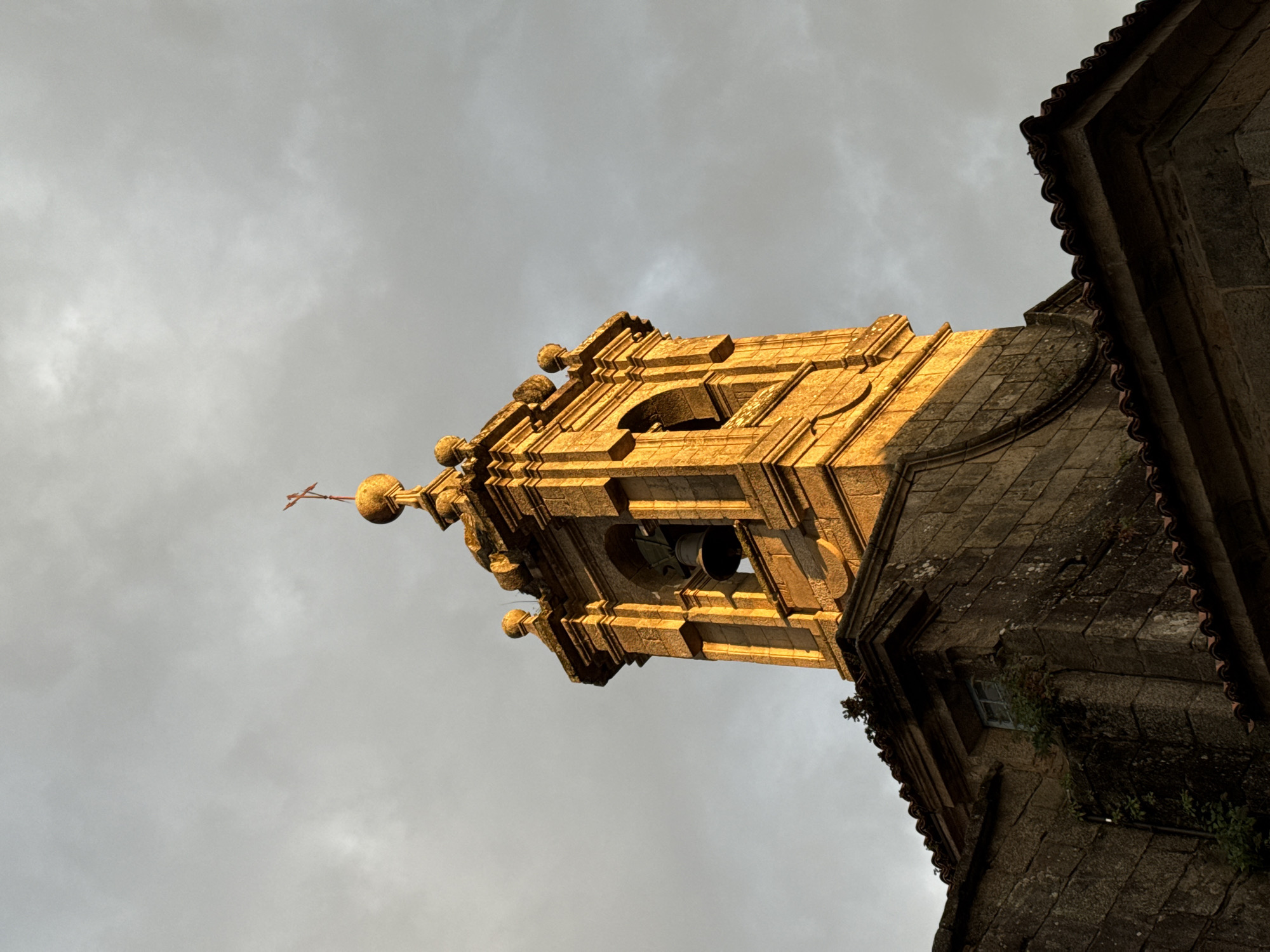 The image captures a close-up, low-angle view of a church bell tower and the adjacent roofline of a stone building, likely located in the city of Santiago De Compostela, Spain. The sky is overcast with dramatic, cloudy textures. The bell tower, constructed of stone, is bathed in the warm, golden light of what appears to be sunset or sunrise, highlighting its ornate architectural details, including arched openings, decorative finials, and a cross at its apex. A bell is visible within one of the tower's openings. The building's roof, covered in red tiles, extends into the foreground, with a portion of the stone wall visible below the tower. Overgrown vines cling to the stonework. The scene is devoid of any people or animals.