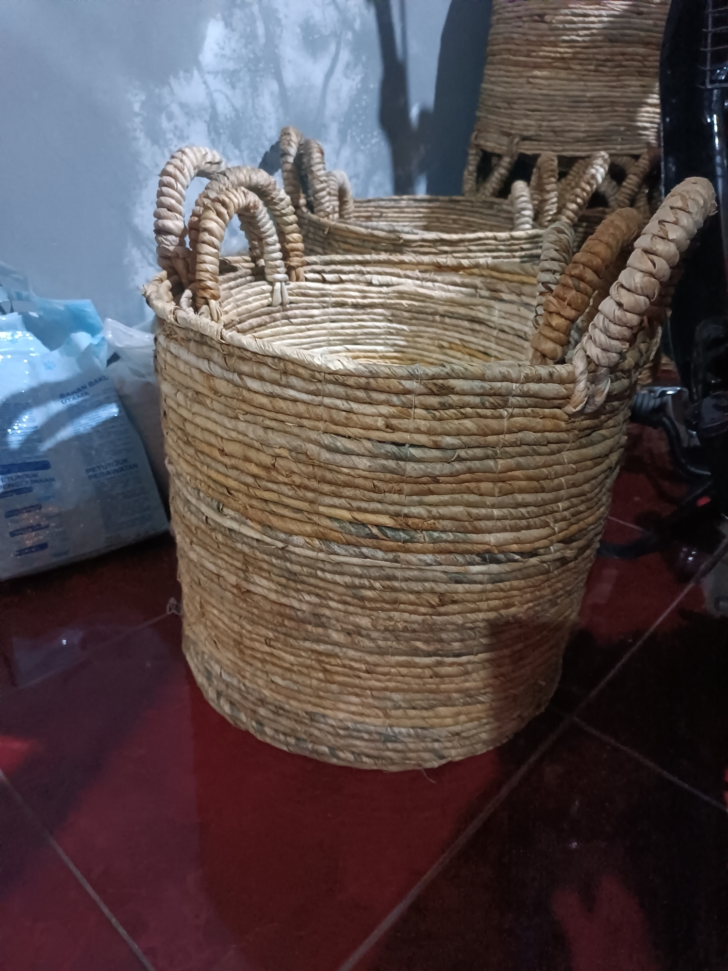 The image shows a close-up of a woven basket with handles, likely made of straw or a similar natural material. The basket is cylindrical and textured with tightly woven strands. Several thick, coiled handles are visible arching over the rim of the basket. In the background, there is a light-colored, possibly textured wall, and to the left, a partially visible blue and white plastic bag with Indonesian text, suggesting it might contain goods. The floor appears to be a dark red, possibly tiled surface. The lighting is dim, casting shadows and highlighting the textures of the basket. There is no indication of the city of Jetis, Indonesia within the image itself; this location information is external to the visual data. The scene appears to be indoors, possibly in a storage area or a room with utilitarian elements. No people are visible in the image.