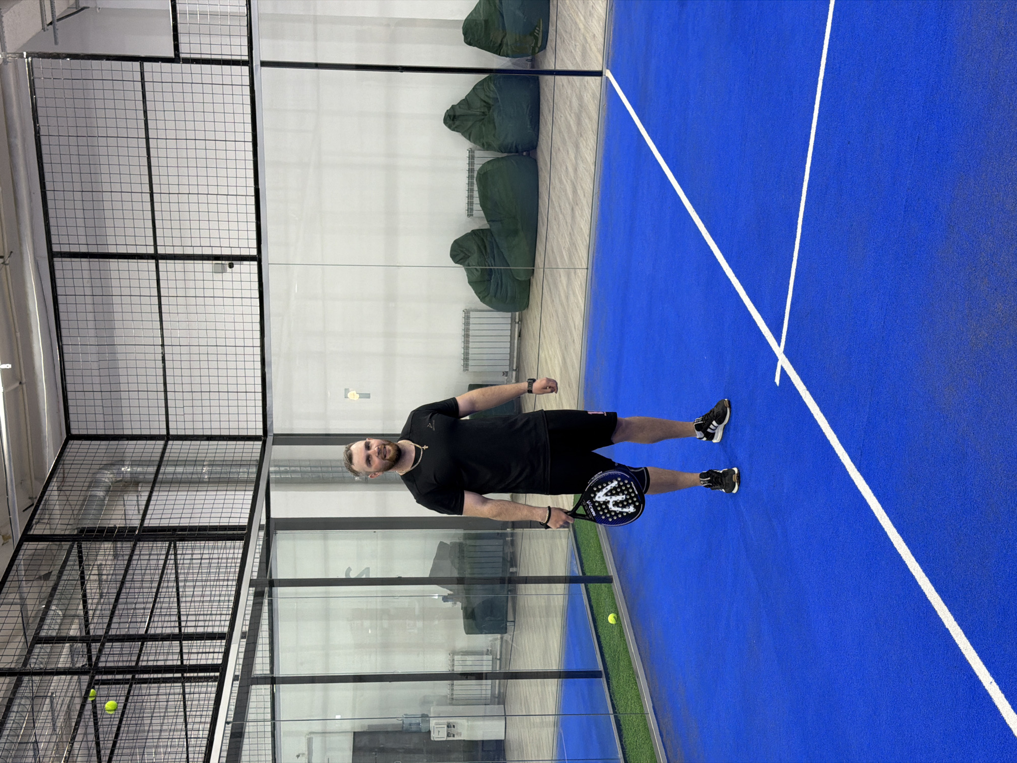A man stands on a blue padel court inside a building with a wire mesh ceiling and glass walls. He is holding a padel racket in his right hand. There is a yellow tennis ball on the floor near the glass wall. Several green beanbag chairs are lined up against the back wall. The scene is likely in an indoor sports facility. The location is unknown.