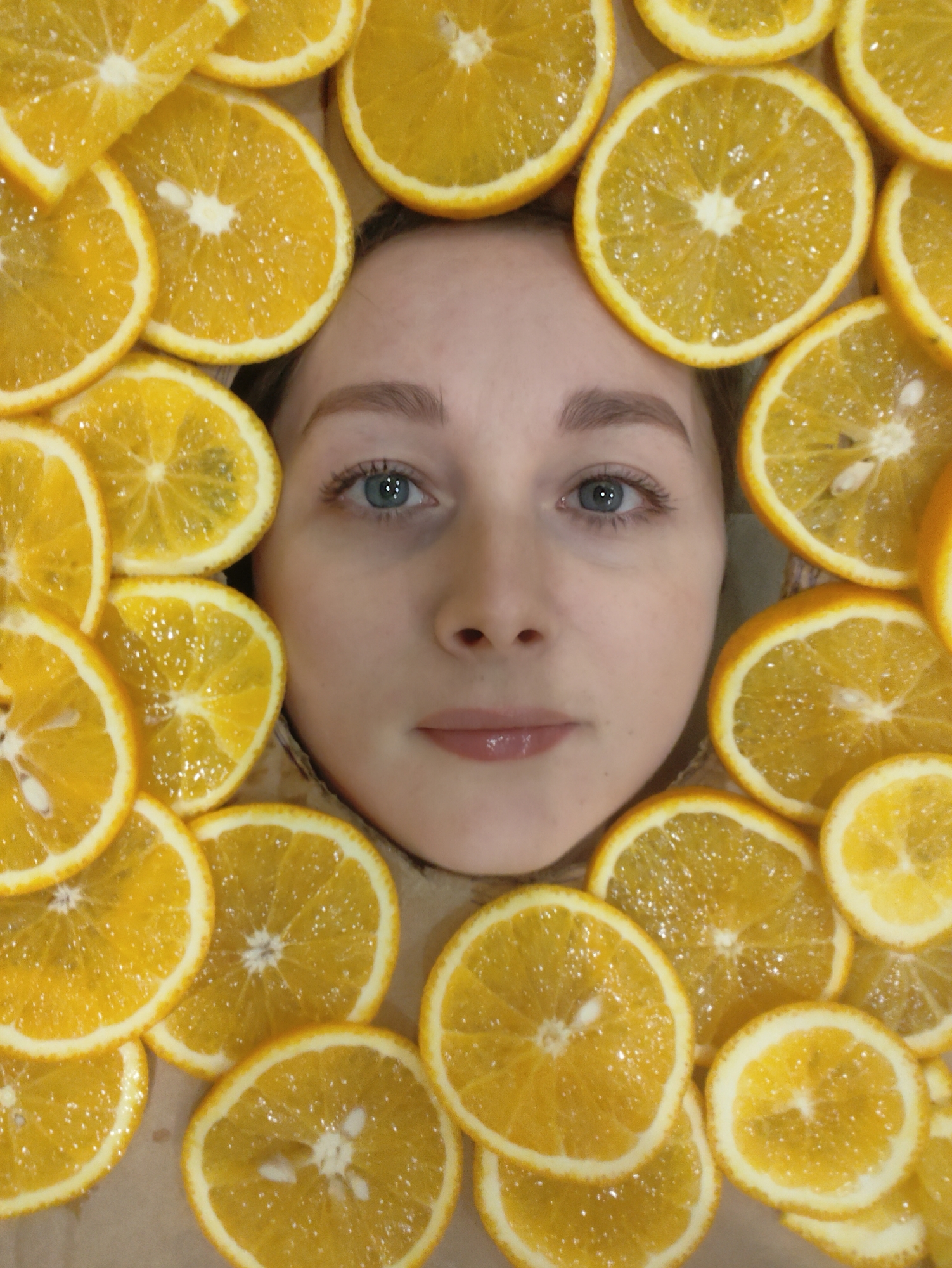 A close-up overhead shot shows a person's face surrounded by orange slices. The person appears to be lying down, with the orange slices arranged around their face to create a frame. The person has light skin, blue eyes, and light eyebrows. The orange slices are bright orange and slightly translucent. The background is a neutral color. The location is Unknown, Unknown.