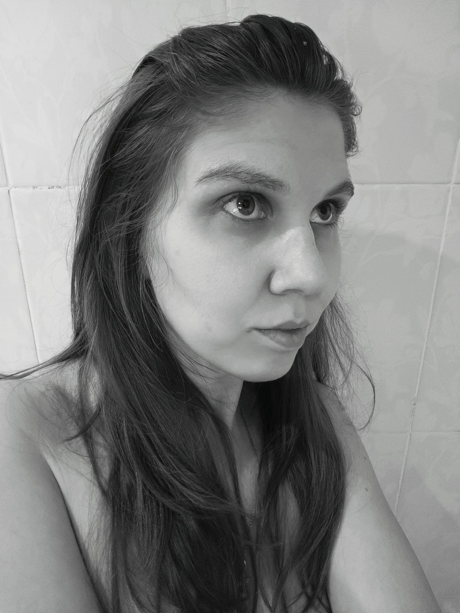 A black and white, eye-level portrait features a young woman looking upwards and to the right with an intense, pensive, or thoughtful expression. Her dark, long hair frames her face, with some strands falling forward and others appearing slicked back or damp near her forehead. Her eyes are wide, reflecting a light source, and her lips are slightly parted. She is shown with bare shoulders, suggesting she is wearing a strapless top or is partially undressed. The background consists of light-colored, possibly tiled walls with visible grout lines, suggesting an indoor setting like a bathroom. The lighting illuminates her face from the front or above, creating highlights and shadows that emphasize the monochromatic tones. No text is visible.