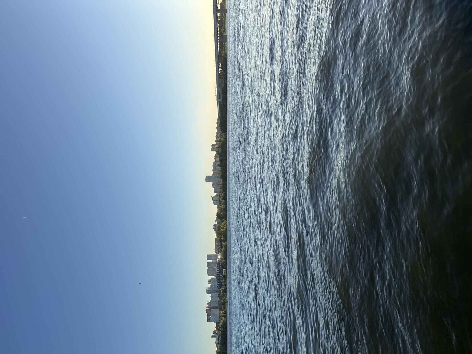 The image depicts a wide view of a river under a clear blue sky. The river occupies the right half of the frame, with its surface rippling, reflecting the sunlight. The left half of the frame shows a distant city skyline along the riverbank, characterized by various buildings and trees. A bridge is visible in the far distance, spanning across the river. The sky transitions from a light yellow hue near the horizon to a deep blue at the top. There are a few small, dark specks in the sky, possibly birds or distant aircraft, and a faint crescent moon shape is visible in the upper left quadrant. The lighting suggests it is either early morning or late afternoon. The scene is serene and tranquil, with no visible people. The location is identified as Novosibirsk, Russia.