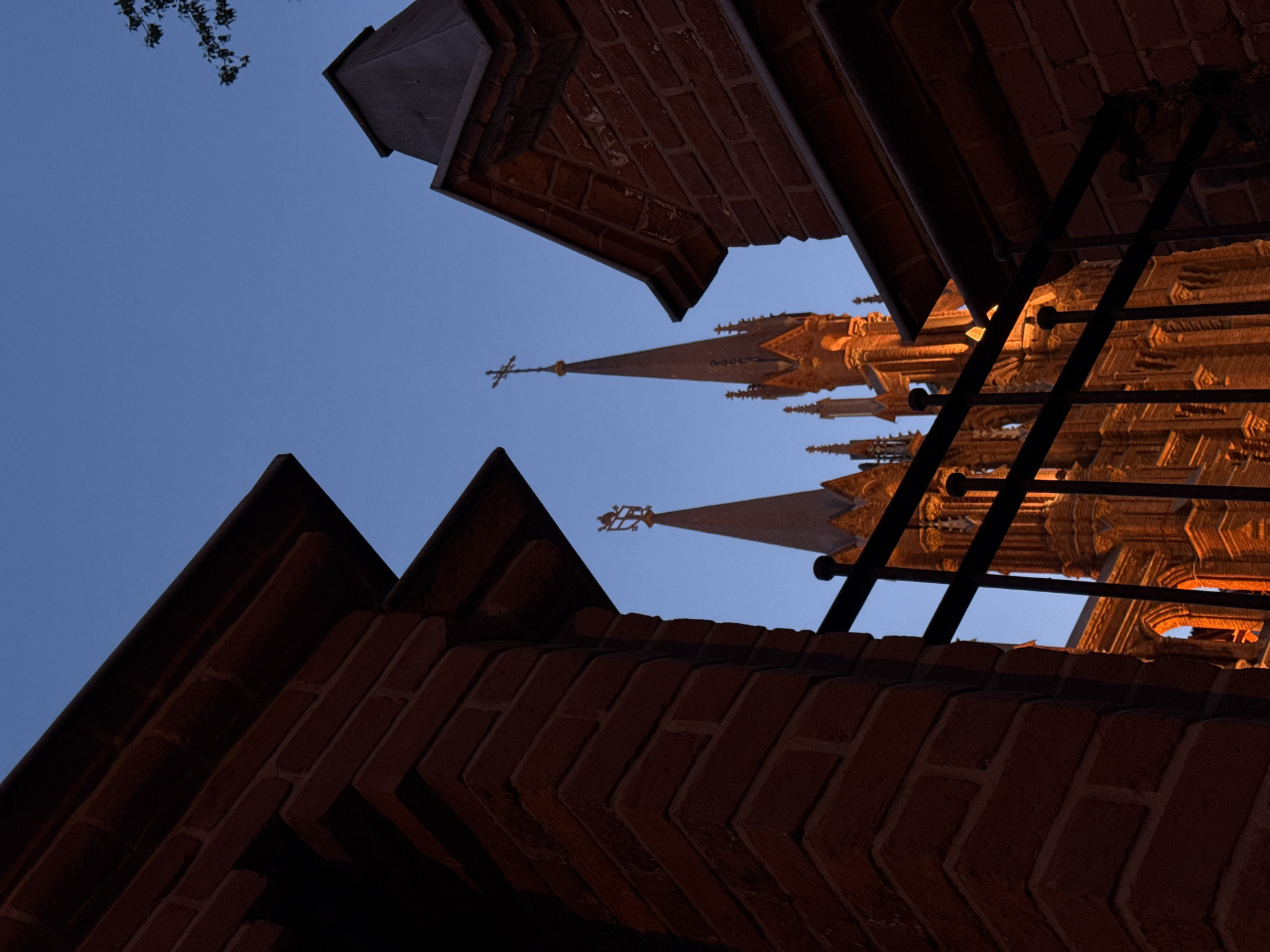 The photograph captures a low-angle perspective of an architectural structure, showcasing brickwork and spires against a twilight sky. The brickwork, primarily in the foreground, is detailed and exhibits a repetitive pattern. In the background, several illuminated spires rise into the sky, adorned with decorative elements including crosses and pinnacles. A metal railing is also visible, partially obscuring the spires. The sky is a gradient of light to dark blue, suggesting dusk or dawn. The building is identified as St. Andrew's Anglican Church in Moscow, Russia.