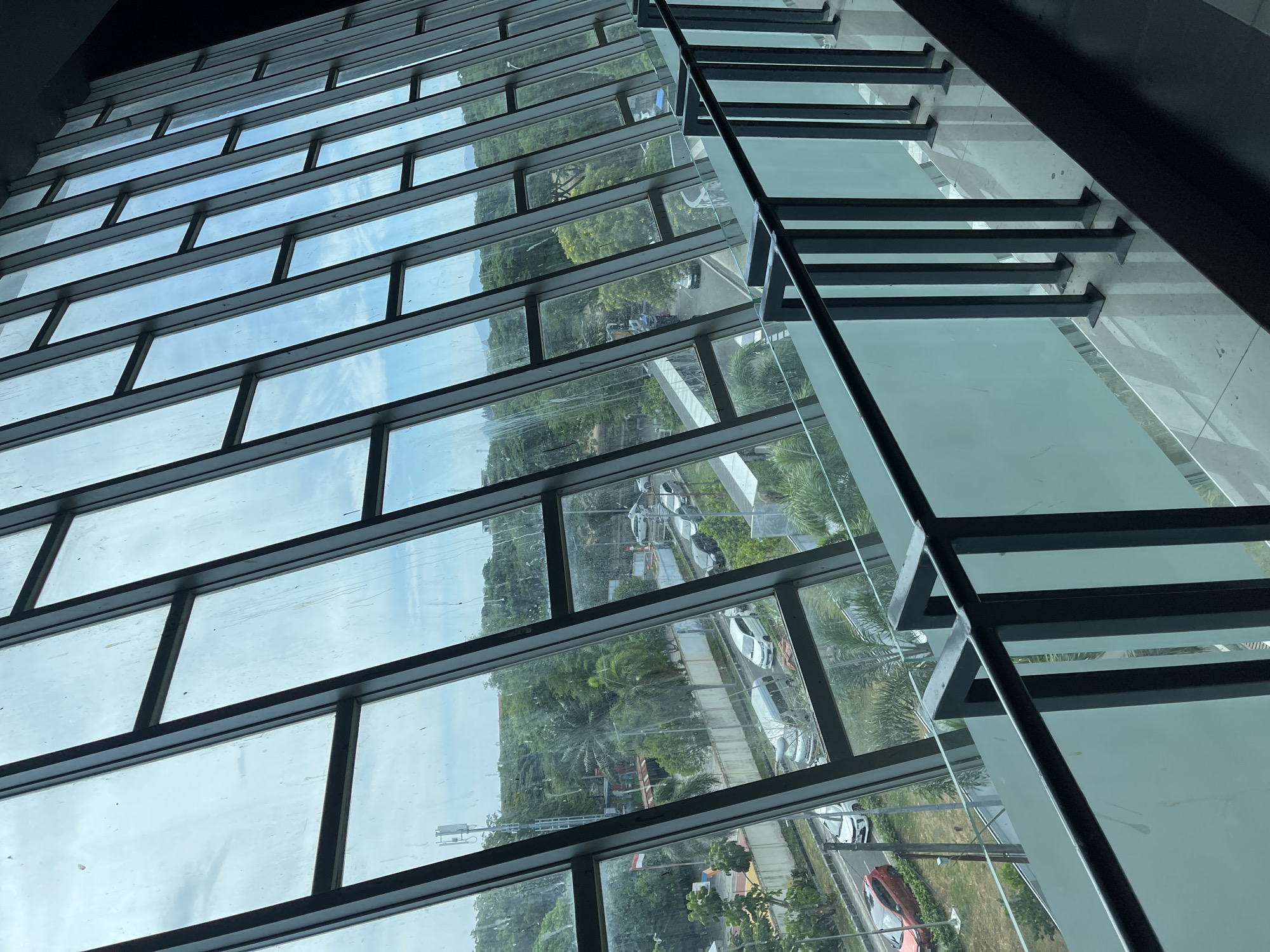 The image is a view through a slanted glass window overlooking a city street in Banda Aceh, Indonesia. The window is framed by dark metal beams and shows a clear, overcast sky. Below the window, a metal railing with frosted glass panels separates the interior from the exterior view.
The exterior scene depicts a street with several vehicles, including cars and what appears to be a van or small bus. Trees and greenery are visible along the roadside and in the background, suggesting a tropical or subtropical environment. A cell tower can be seen in the distance. The architecture of the building, indicated by the slanted windows and interior railing, suggests it might be a modern commercial or public building. The road appears to be asphalt, and there are sidewalks or pedestrian areas visible. The presence of an Indonesian flag in the distance further confirms the location. The overall impression is of an urban landscape with a good amount of green space.