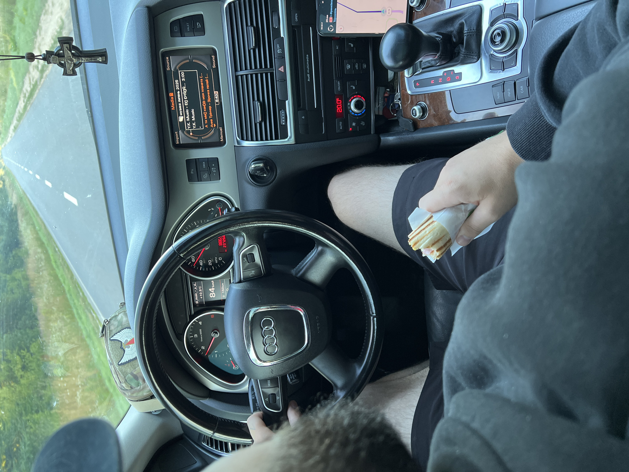 The image depicts the interior of a car, likely an Audi Q7 based on the steering wheel and dashboard design, as it travels down a paved road. The road is bordered by green foliage, suggesting a rural or semi-rural setting. A male driver, with a beard visible, is holding the steering wheel with his left hand and a sandwich with his right. The sandwich appears to be a wrap with ham and cheese.
The car's dashboard is illuminated, showing the current speed at 84 km/h and the outside temperature at +10.5°C. The infotainment system displays "музика люмень 2025" (music lumen 2025) and song titles in Cyrillic. A car accessory, possibly a rosary or pendant, hangs from the rearview mirror. A camouflage patterned pouch with a red and white emblem is visible on the dashboard.
The central console features the gear selector, climate control, and navigation buttons. A smartphone is mounted on the console, displaying a navigation interface with the destination "Тернопіль" (Ternopil) and a distance of 25 km. The driver is wearing a dark hoodie and black shorts. The overall scene suggests a journey is in progress. The location mentioned, Darakhiv, Ukraine, aligns with the presence of Cyrillic text on the infotainment system and the navigation destination.