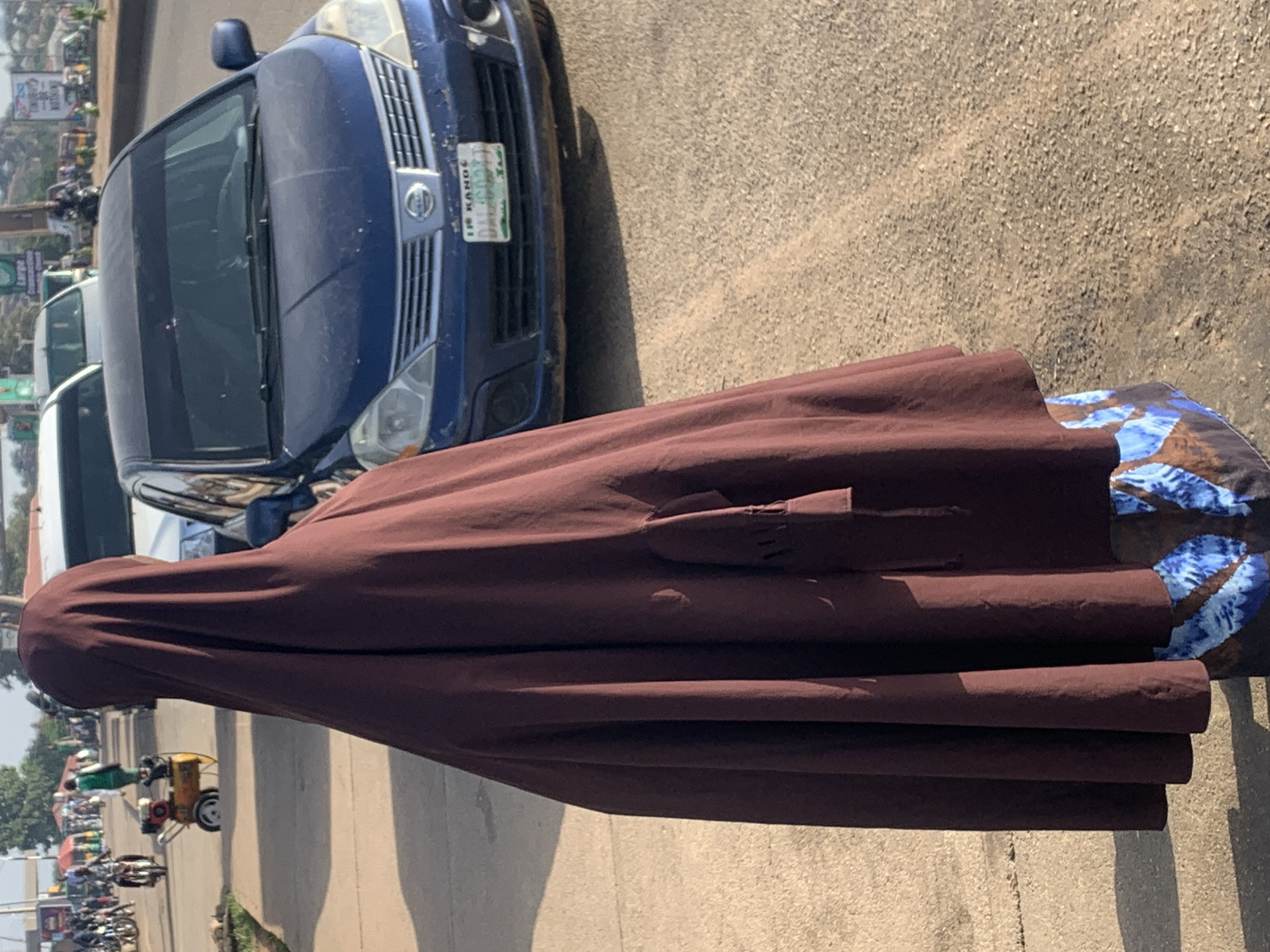 A person in a dark maroon hijab and patterned skirt stands on a dusty roadside in Jos, Nigeria, facing away from the camera. Behind the person, a dark blue Nissan sedan is parked on the road, with its license plate visible. In the background, various vehicles, including motorcycles and what appears to be a generator on a cart, are visible. The scene suggests a bustling street or market area.