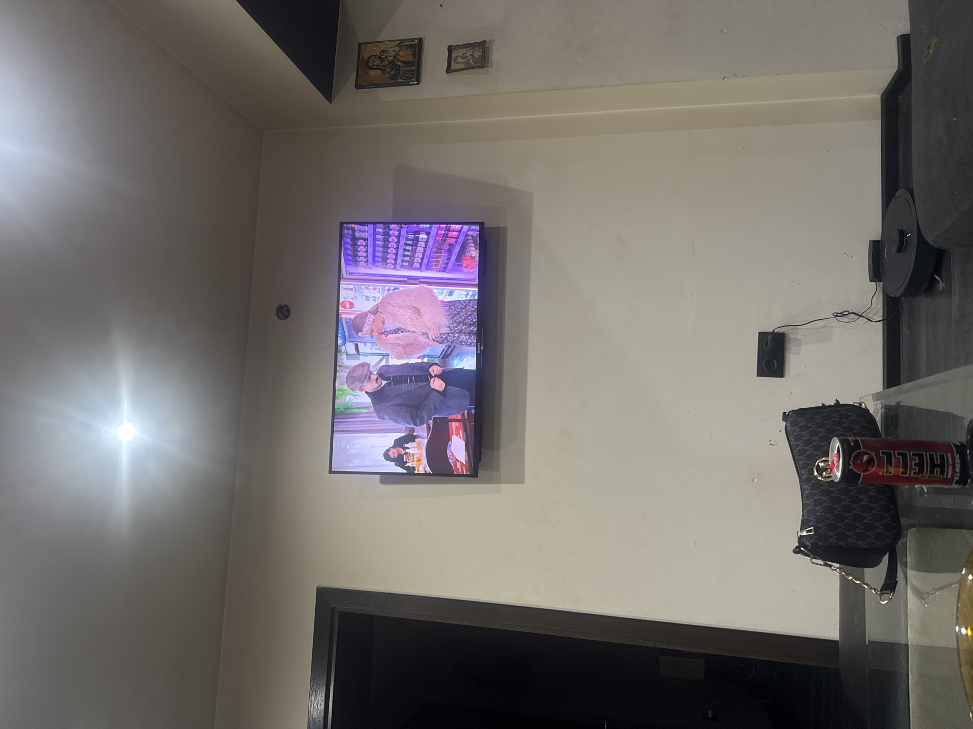 An indoor room, likely a living area, is depicted with light-colored walls and a dark door frame visible at the bottom. A flat-screen television is mounted on one wall, displaying a program. On the TV screen, two individuals are visible: an upper figure, a woman wearing a pink, furry top or jacket and a headband, and below her, a man dressed in a plaid jacket and a flat cap. The background of the TV program appears to be a retail environment, possibly a supermarket with shelves.
Above the television, two framed pictures or icons are hung high on the wall. To the right of the TV, a black speaker or power adapter is affixed to the wall, with electrical wires extending from it. A bright light source or reflection is prominent on the wall to the left of the TV.
On the right side of the room, a dark, vertical structure is partially visible, and a glass table is present. Resting on the table are a black purse adorned with a repeating "M" pattern and a red energy drink can.
The activity taking place is the viewing of television. The energy drink can prominently displays the brand name "HELL" (appearing upside down in the current image orientation), and a small logo (possibly "Y" or "V") is visible on the bottom left of the TV screen. The time of day appears to be indoors under artificial light. No specific location cues for Dolna Gradeshnitsa, Bulgaria, are visually present within the image.