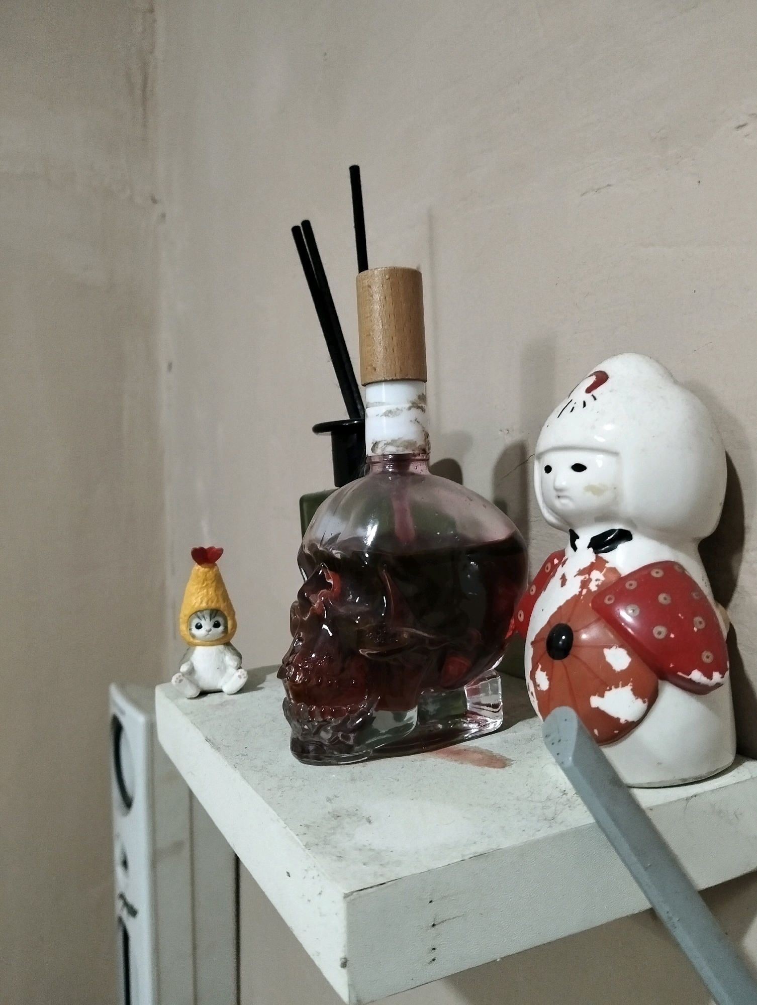 This is a close-up, eye-level, indoor shot of a shelf in Patrol, Indonesia. The shelf, made of white painted wood, has several decorative objects. To the left is a small, white cat figurine wearing a yellow hat with a red tip. Next to it is a clear glass bottle shaped like a skull, filled with a dark red liquid. Three black diffuser reeds are inserted into the bottle. To the right of the skull bottle is a white and red ceramic doll with a large, round white headdress and a red and white patterned garment. A grey object, possibly a ruler or a piece of molding, is placed diagonally across the front of the shelf. The background is a textured, light-colored wall. A portion of a white appliance with vents is visible to the left of the shelf. The lighting is moderate, creating soft shadows.