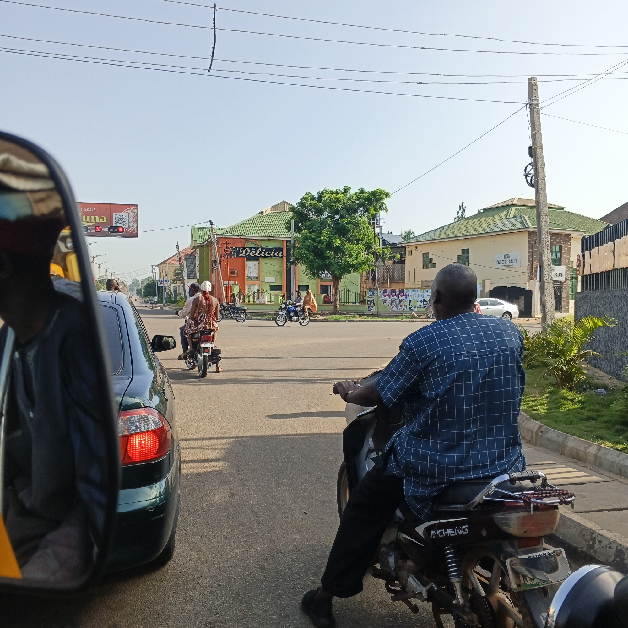 The image depicts a street scene in Kabala, Nigeria, under a clear sky. In the foreground, the back of a motorcycle is visible, with a person in a blue checkered shirt and black pants riding it. The motorcycle has a license plate that reads "KADUNA". To the left, a dark green car is seen from the side, with its red taillight illuminated. In the middle ground, several other motorcycles and their riders are present, some stopped at what appears to be an intersection. One motorcycle carries two people, a man and a woman wearing a patterned dress.
Further in the background, buildings with green roofs are visible. One building has a sign that says "Delicia" in colorful lettering, with a smaller sign below that appears to say "BAKE HUT". A large tree stands in front of this building. Power lines are strung across the sky, supported by concrete poles. The road surface is asphalt, and there is a sidewalk with green grass and a curb. The overall impression is a typical day in a Nigerian city with moderate traffic.