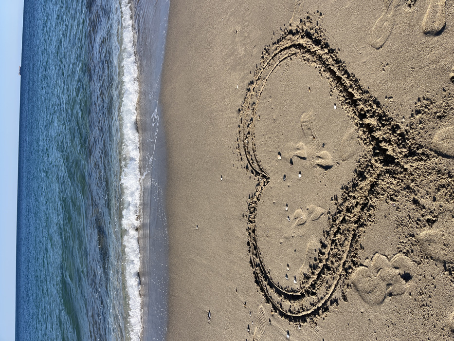 The scene depicts a sunny day on a sandy beach in Alanya, Turkey, facing the Mediterranean Sea. The foreground features light brown, fine-grained sand where a large heart shape has been drawn, deeply etched into the surface. Several indistinct footprints are visible within and around the heart, along with scattered small, light-colored shells or pebbles. The midground shows the shoreline where a small, white-crested wave is actively breaking, creating a line of foam and wet sand. The background consists of the deep blue-green waters of the Mediterranean Sea, transitioning to a clear, light blue sky above the horizon. A dark-colored vessel, likely a cargo ship, is visible as a small silhouette on the distant horizon, slightly right of center. There are no visible individuals present in the frame.