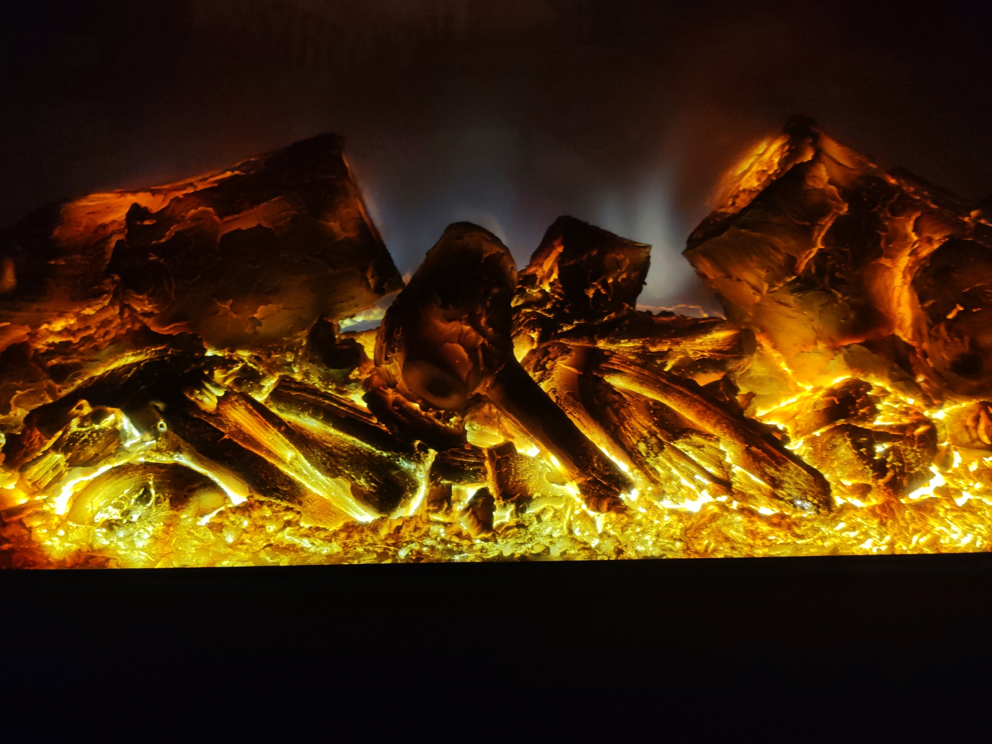 The image displays a close-up view of an artificial fireplace, featuring logs that glow with warm, orange and yellow light. The logs appear to be made of a material designed to mimic burnt wood, with textured surfaces and irregular shapes. Some logs are piled on top of each other, creating depth and dimension. The intense light emanating from the logs casts a warm glow, illuminating the surrounding simulated embers and the darker background. Wisps of blue and white light, suggesting flames or smoke, are visible in the upper portion of the scene, adding to the illusion of a real fire. The bottom of the image is dominated by a dark, undefined area, likely representing the base of the fireplace or the surrounding darkness. There is no visible text in the image. The scene evokes a sense of warmth and coziness, typically associated with a fireplace. The lighting and texture suggest the image was taken in a dimly lit indoor environment, with the artificial fire being the primary light source. The overall impression is one of a realistic imitation of a crackling fire.