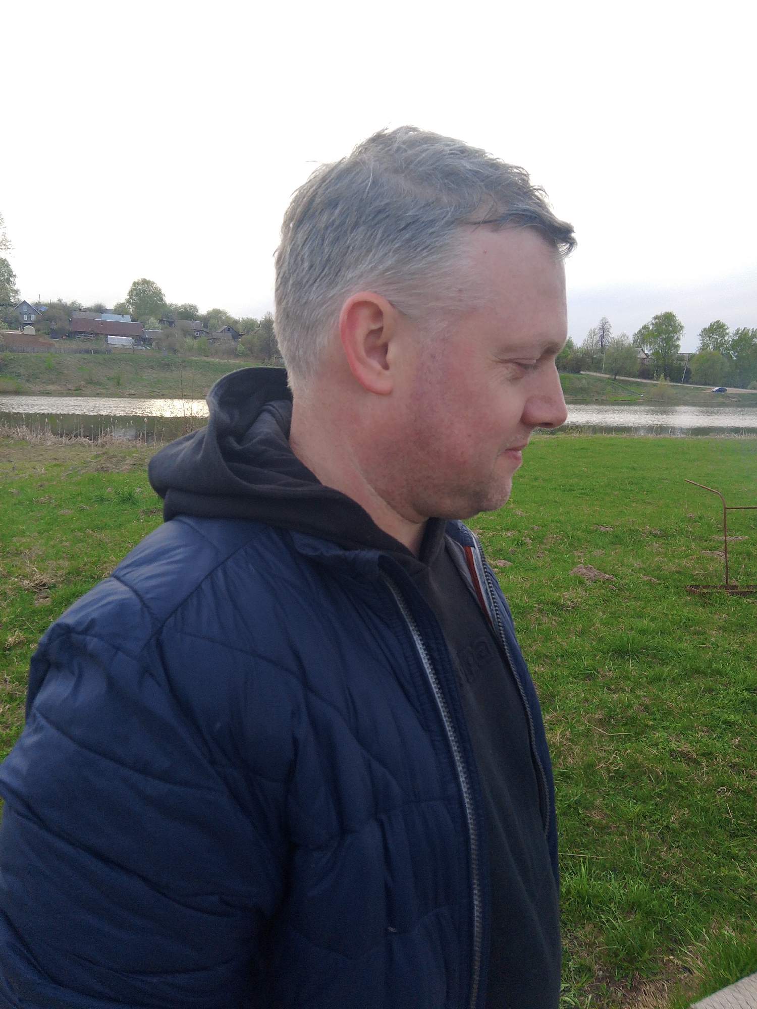 A middle-aged man is standing outdoors, likely in a grassy area near a body of water. He is wearing a dark blue quilted jacket with a black hoodie underneath. The man has short, graying hair and is facing to his right. In the background, there is a body of water and trees. Beyond that, one can see some buildings, possibly indicating a settlement or village. While not visible in the main picture, there is a crop hint indicating the city of Фоки, Russia.