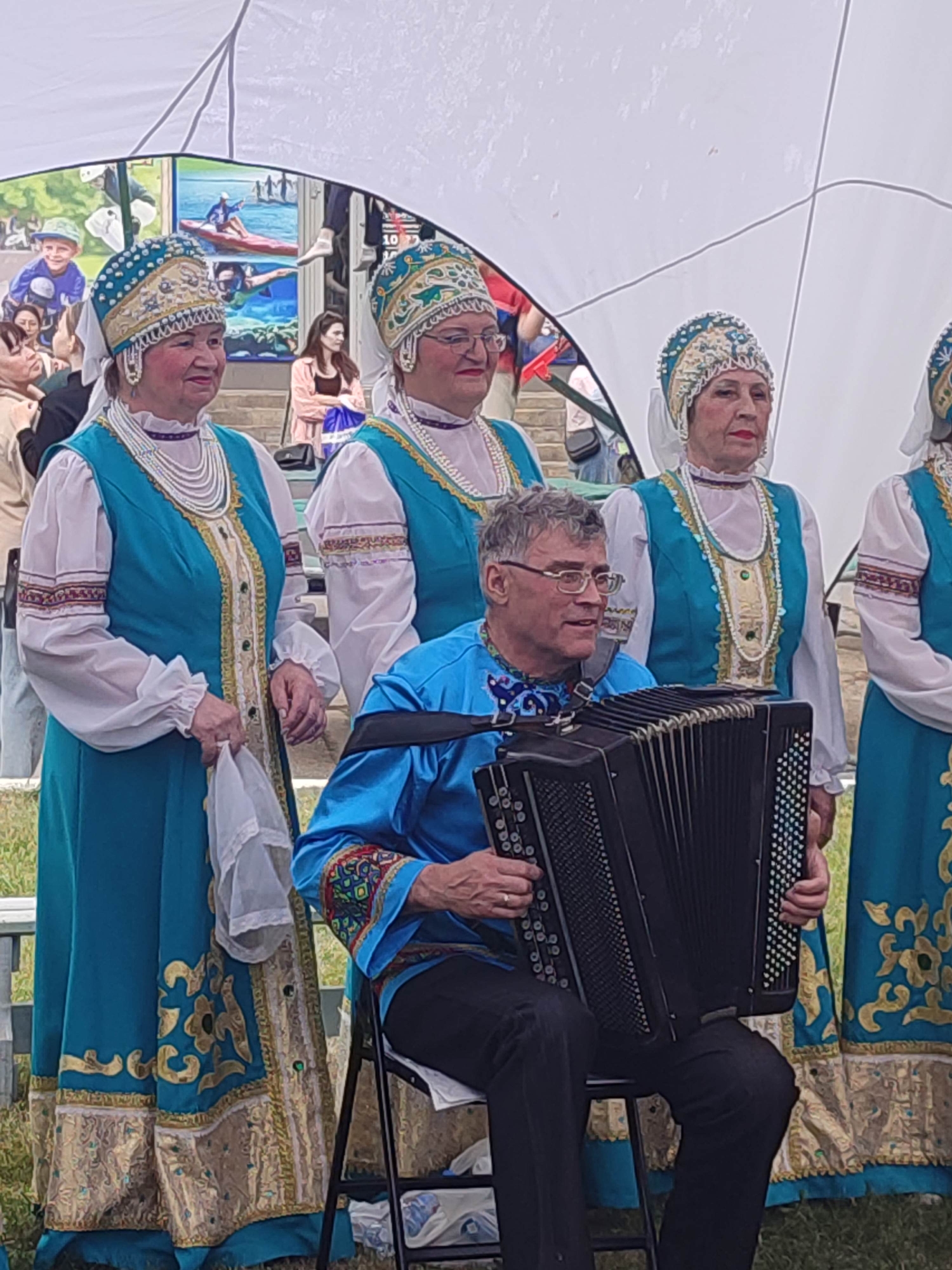 A man plays an accordion while standing with a group of women in traditional attire. The man is seated and wearing a blue shirt with embroidered details on the collar and sleeves. The women are standing and wearing matching teal vests over white blouses, with ornate headpieces, pearl necklaces, and heavily embroidered skirts. The background features a white tent or canopy, stairs, and a mural depicting people engaged in water activities. The setting appears to be outdoors, possibly at a festival or cultural event.