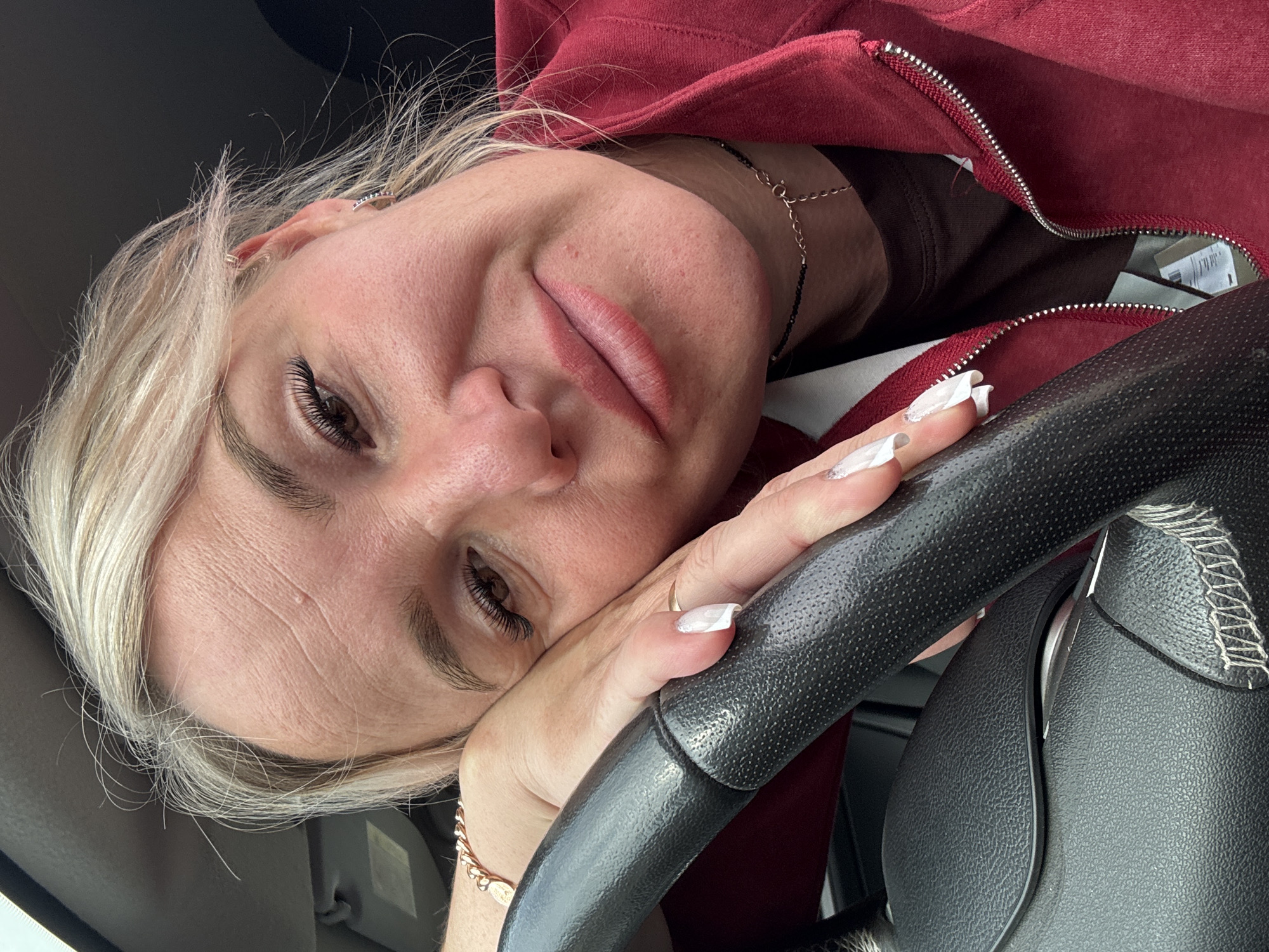 A close-up, waist-up shot features a woman with blonde hair, resting her head on her left hand, which is placed on a black steering wheel. She is looking towards the camera with a slight smile. The woman is wearing a dark brown shirt and a red zip-up jacket. Her left hand displays long, white acrylic nails. A delicate gold necklace is visible around her neck. The background is blurred and appears to be the interior of a car. The scene is likely set within the city of Chastyye, Russia, as indicated by the provided information.