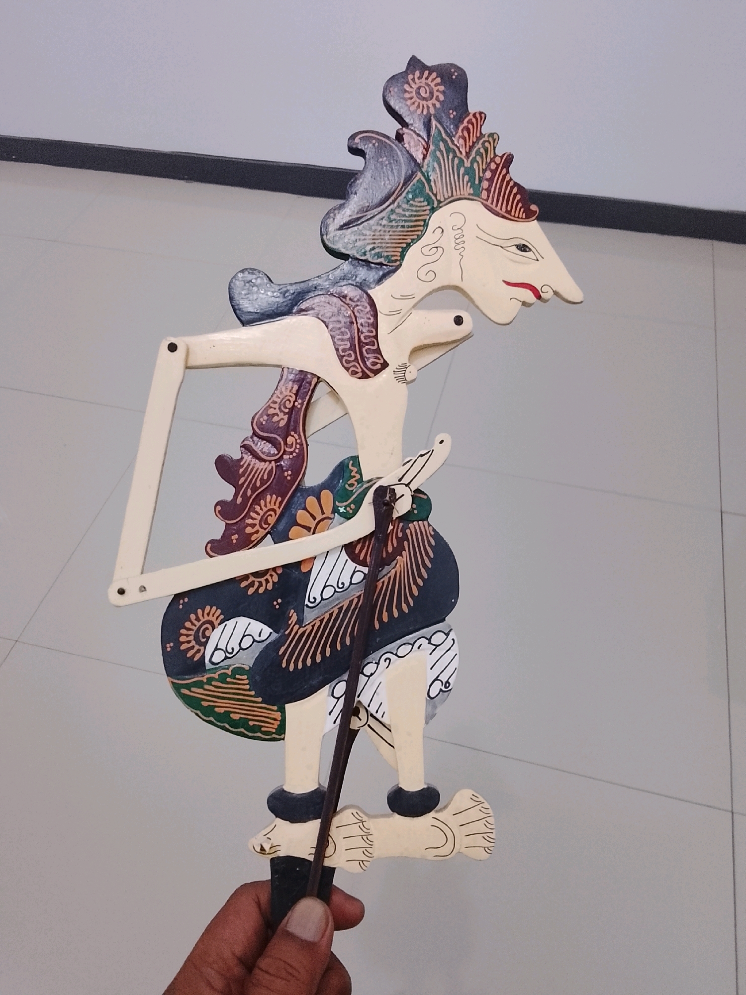 In the city of Slawi, Indonesia, a hand is holding a traditional Wayang Golek puppet. The puppet is made of wood and painted with intricate details. It depicts a character with a stylized face, elaborate headdress, and a richly decorated costume. The puppet is held upright by a wooden stick, and a framework extends from its body, likely for manipulation. The background consists of a tiled floor and a plain wall, suggesting an indoor setting.