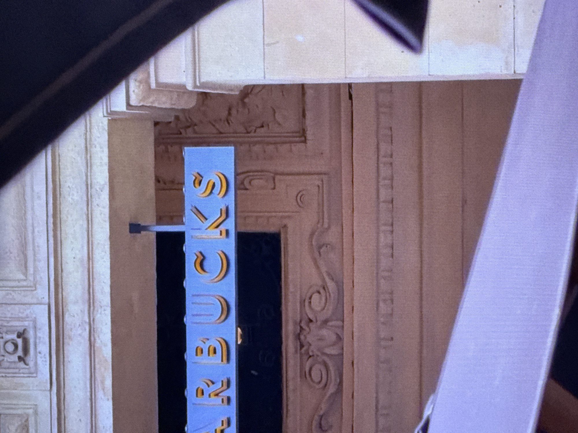 The image shows a partial view of a Starbucks sign mounted on a building with classical architectural elements. The sign itself is blue with raised golden lettering that appears to spell out "STARBUCKS." The building facade features decorative molding and stonework. A dark, angled object, possibly part of a vehicle or structure, partially obstructs the view on the right side of the frame. The overall impression is of an urban environment, though no specific city or location is identifiable from the image alone.
