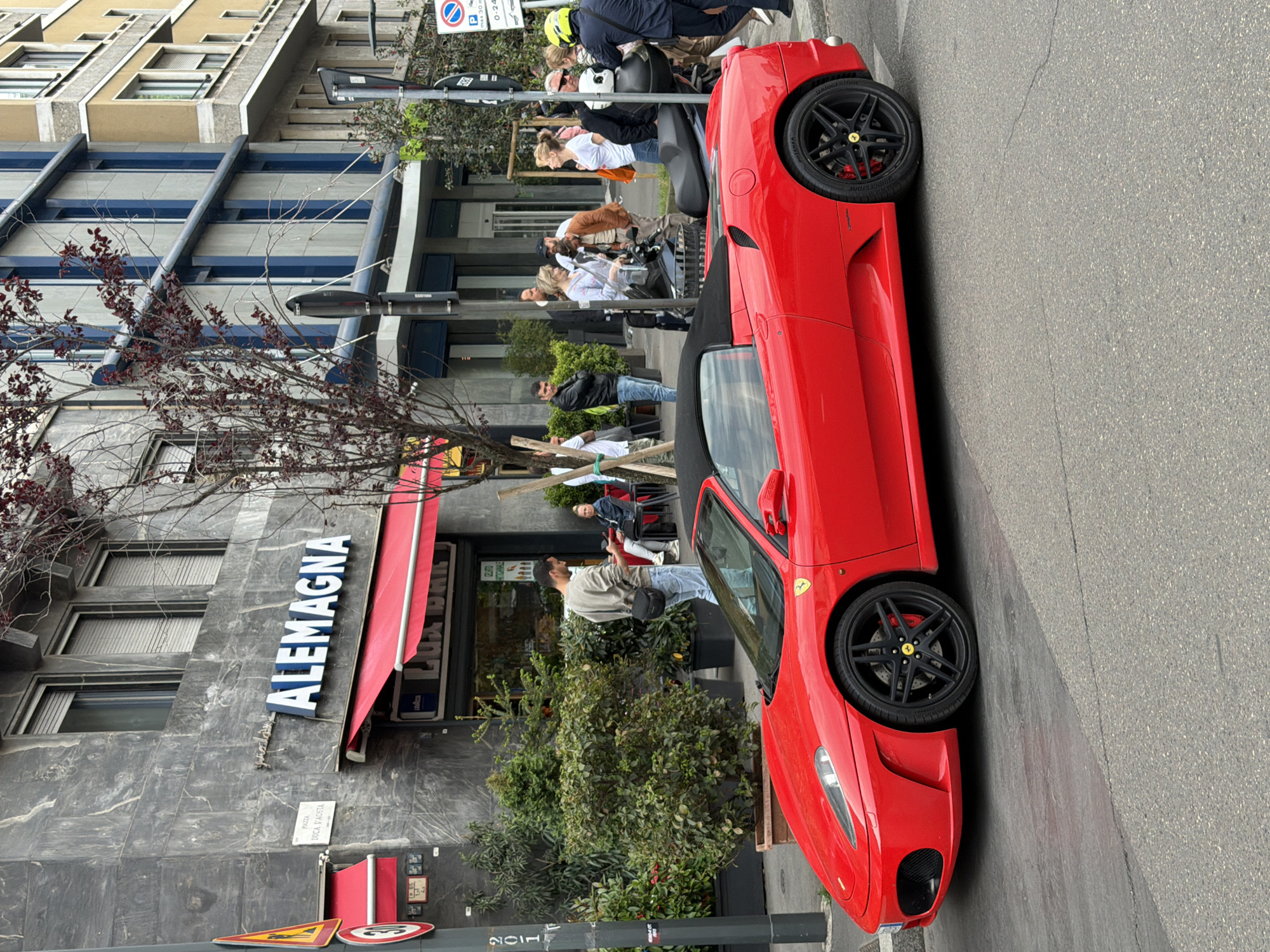 A vibrant urban street scene in Milan, Italy, features a sleek red Ferrari F430 Spider parked prominently on the side of an asphalt road. The luxury convertible has black multi-spoke wheels with yellow Ferrari center caps and red brake calipers, with "BRIDGESTONE" visible on the tire sidewalls.
To the left of the car, a multi-story building with a red awning houses a cafe or shop. A large sign on the building clearly reads "ALEMAGNA" in white letters with a blue outline, and "LUS BAR" (likely "LUX BAR") with "LAVAZZA" branding is visible on the awning. In front of this establishment are green bushes and sparse trees with reddish leaves. Several people are gathered in this area; some appear to be standing or walking, while others might be seated at outdoor tables.
Further into the background, other buildings with modern architectural elements, including prominent horizontal windows and balconies, line the street. A variety of people are visible throughout the scene, some walking, others on scooters or motorbikes. One person wears a distinctive yellow helmet.
Various street furniture and signs are present: a street light pole, multiple traffic signs (including a circular blue "no parking" sign with "0-24" and "max 30 min," a triangular yellow warning sign, and a circular red "30" speed limit sign). A white street sign on the building clearly indicates "PIAZZA DUCA D'AOSTA," confirming the specific location in Milan.
The time of day appears to be daytime, with soft, diffused lighting suggesting an overcast sky. The overall impression is a bustling yet relaxed city environment.