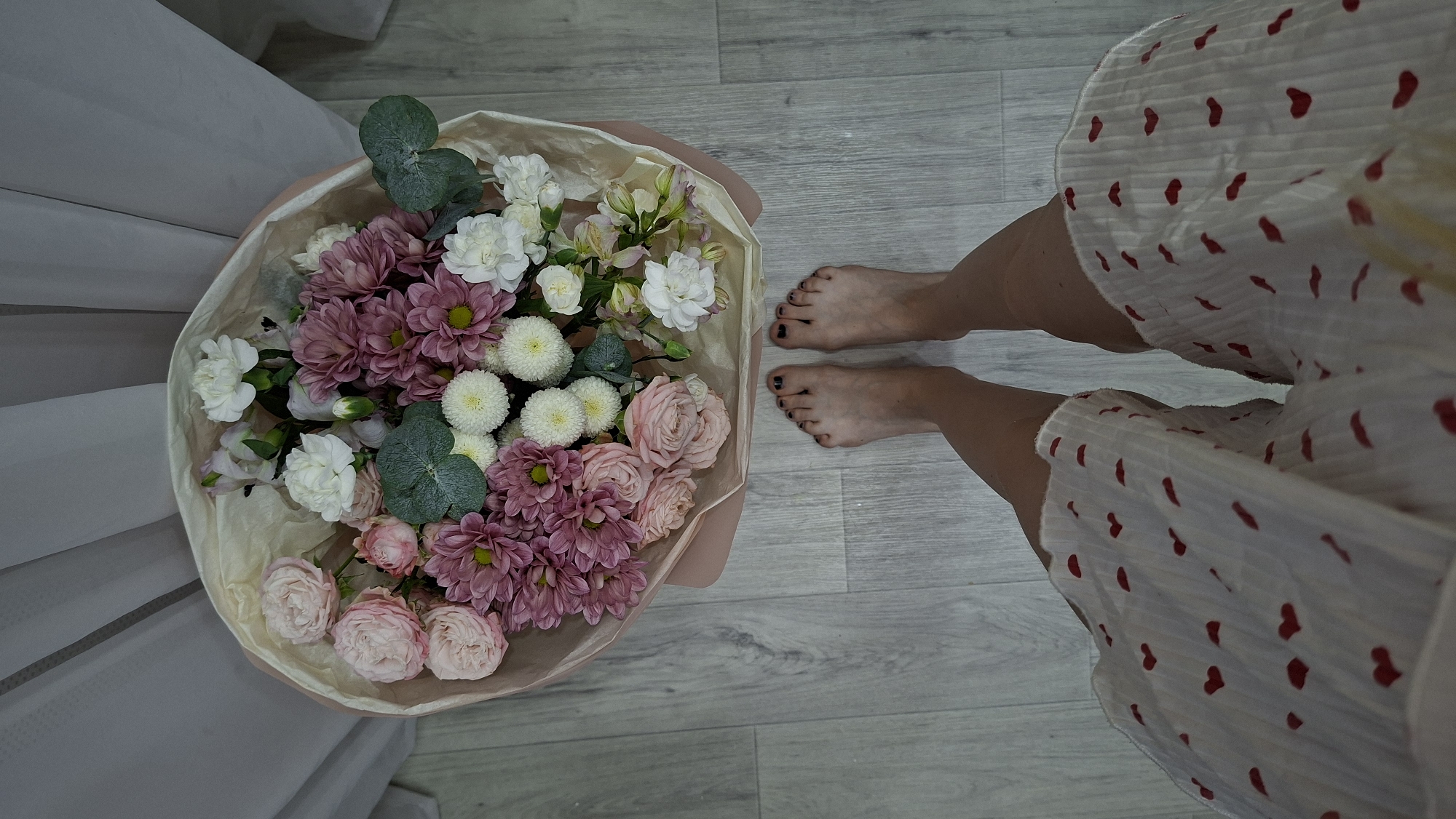 The image shows a top-down view of a bouquet of flowers and a person's bare feet. The bouquet is composed of pink roses, purple dahlias, white carnations, and small white chrysanthemums, wrapped in light-colored paper. The person's feet, with black nail polish, are positioned on a light wooden floor. The lower half of the image shows the person wearing a white skirt with a pattern of red hearts. In the background, light-colored curtains are visible. The scene is indoors. There is no indication of the city of Chaikovsky, Russia.