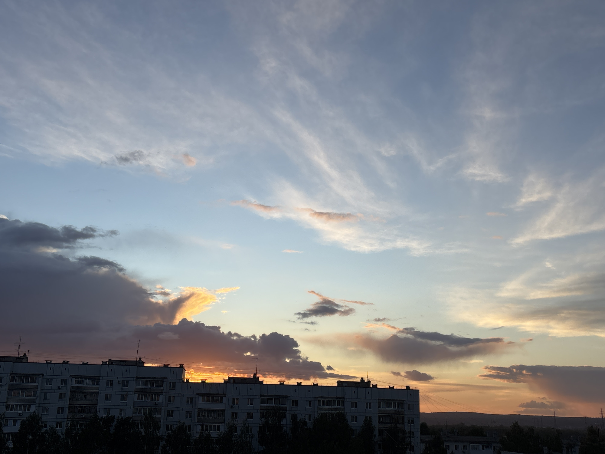 The image shows a sunset over a building in Chaikovsky, Russia. The sky is a mix of blue, white, and orange, with various cloud formations. The building is in the foreground, with trees lining the bottom of the frame. The horizon is visible in the distance.