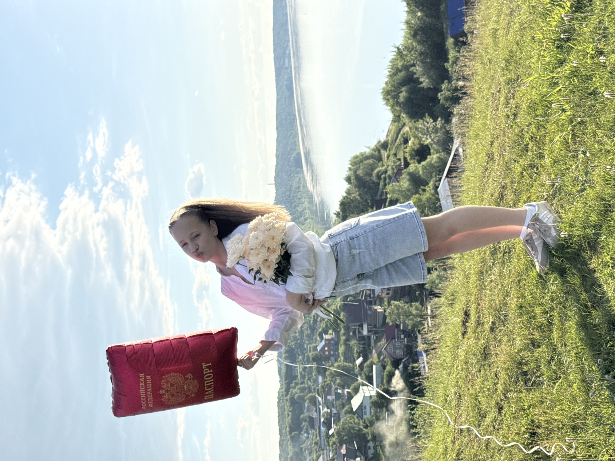 The image depicts a young, light-skinned female individual standing on a sunlit grassy hillside, located in or near the city of Ольховка, Russia. She holds a red foil balloon shaped like a Russian passport in her left hand, which features the text "РОССИЙСКАЯ ФЕДЕРАЦИЯ" (Russian Federation) and "ПАСПОРТ" (Passport) along with the Russian double-headed eagle emblem. A white string is attached to the balloon. In her right hand, she carries a bouquet of light-colored, possibly cream-colored, flowers. She is dressed in a light-colored long-sleeved shirt, light-wash denim shorts, white socks, and light-toned sneakers. Her head is tilted backward and to her left, with her lips pursed.
In the mid-ground, below the hillside, a village with numerous houses of varying colors and architectural styles is visible, interspersed with dense green trees and foliage. Wisps of smoke are observable rising from at least one point within the village. The far background reveals a wide, calm body of water, likely a river, reflecting the bright blue sky. The opposite bank of the river is covered by an extensive forest, and a tall, slender structure is faintly visible on the distant horizon to the right. The sky is clear with scattered white cumulus clouds. The scene is illuminated by natural sunlight, casting distinct shadows on the grass.
