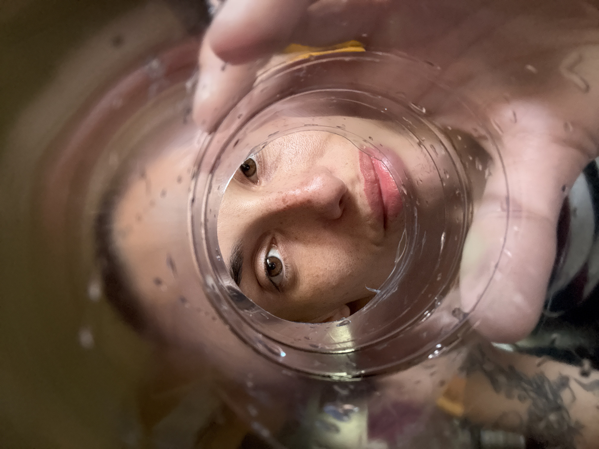 An intimate and distorted close-up shot captures a person's face viewed from an unusual perspective: through the bottom of a clear, cylindrical container, possibly a glass or bottle. The central focus is on the person's right eye, nose, and mouth, which appear magnified and warped by the container's surface. The eye is brown, and the skin exhibits freckles. The lips are visible, tinted with a pinkish-red hue, suggesting lipstick or natural color.
Part of a hand, with fingers and palm, is visible holding the container, extending into the upper and right portions of the frame. A blurred tattoo can be seen on the lower right, on what appears to be the person's arm or hand. The container itself shows subtle rings and numerous small water droplets, adding to the visual texture and suggesting it might contain a liquid. The background is heavily blurred and out of focus, creating a shallow depth of field that emphasizes the person's face. The lighting is soft and even, typical of indoor conditions. There is no visible text in the image. The scene is set in Pleven, Bulgaria.